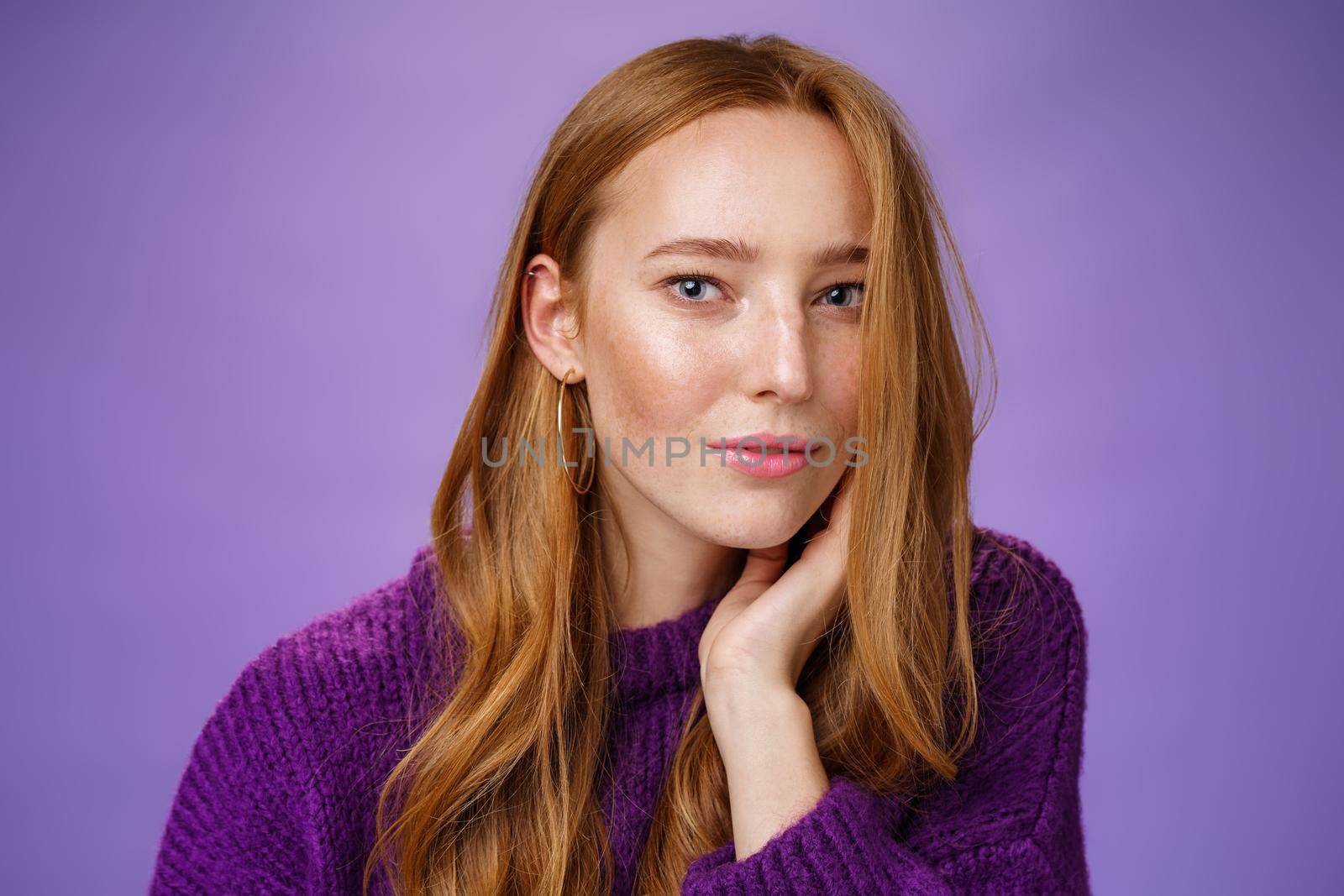Sensual and romantic woman with red hair and freckles touching cheek gently as curls dropping on half face gazing charmed and flirty at camera, smiling being tender and coquettish over purple wall.