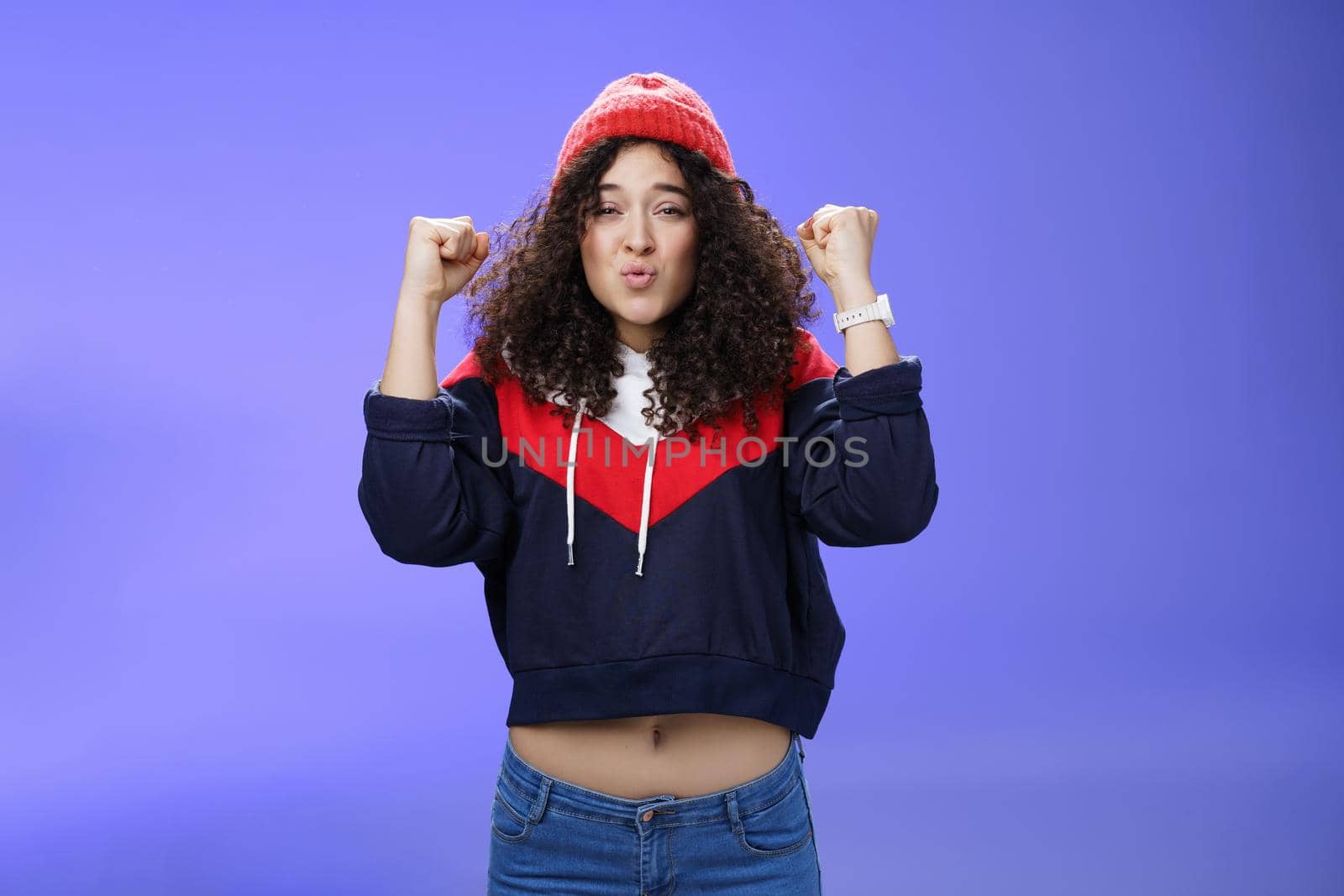 Charming young 20s girl with curly hair in warm beanie raising hands as belly showing under sweatshirt triumphing, celebrating awesome win folding lips from joy and excitement over blue background by Benzoix