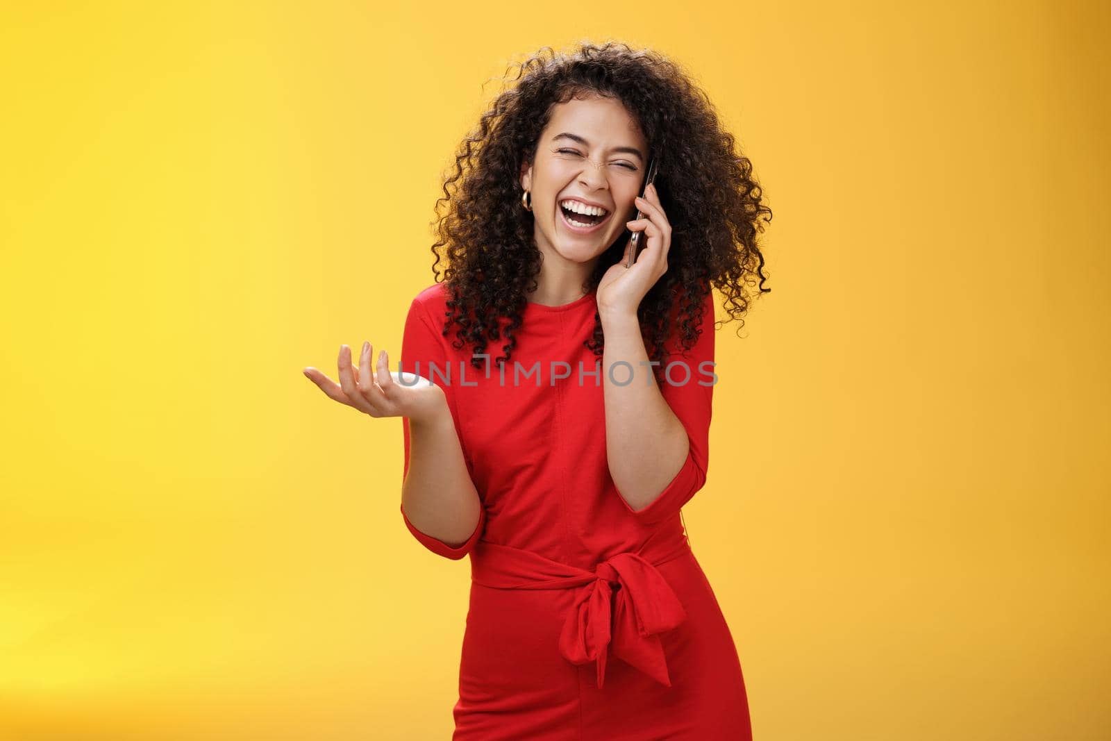 Portrait of joyful charming european female in red dress with curly hair laughing out loud as gossiping with friend via smartphone close eyes as giggling gesturing, hearing joke through mobile phone.