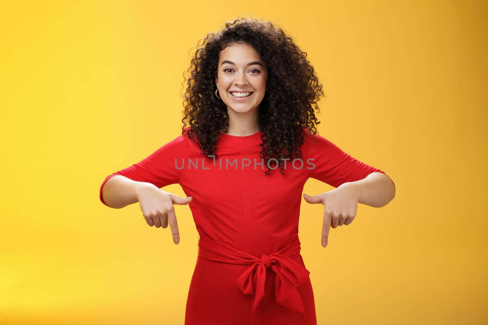 Studio shot of pleasant smiling young curly-haired woman in red dress pointing down with index fingers as promoting awesome event smiling broadly suggesting try or buy over yellow wall.