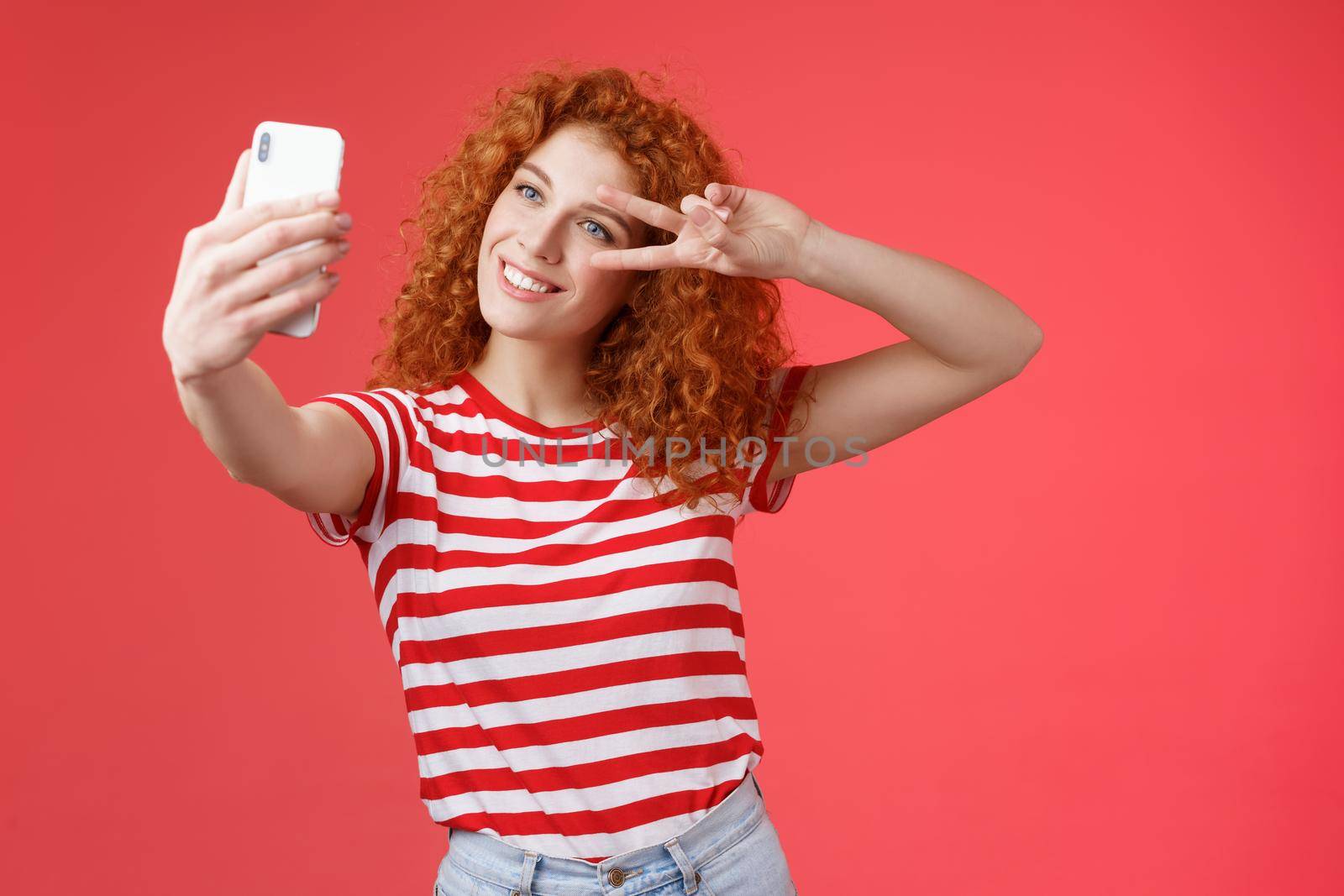 Hey followers how your summer holidays. Cheerful self-assured stylish fashionable redhead curly girl strike pose show peace victory gesture look smartphone screen taking selfie red background.