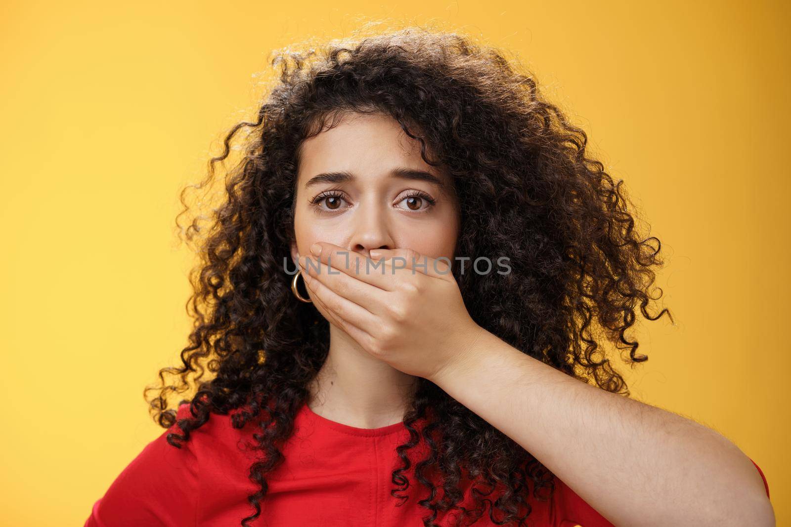 Close-up shot of concerned and insecure troubled woman keep silent scare of telling anyone her problem covering mouth with palm not to scream or slip word looking sad and sorrow over yellow background.
