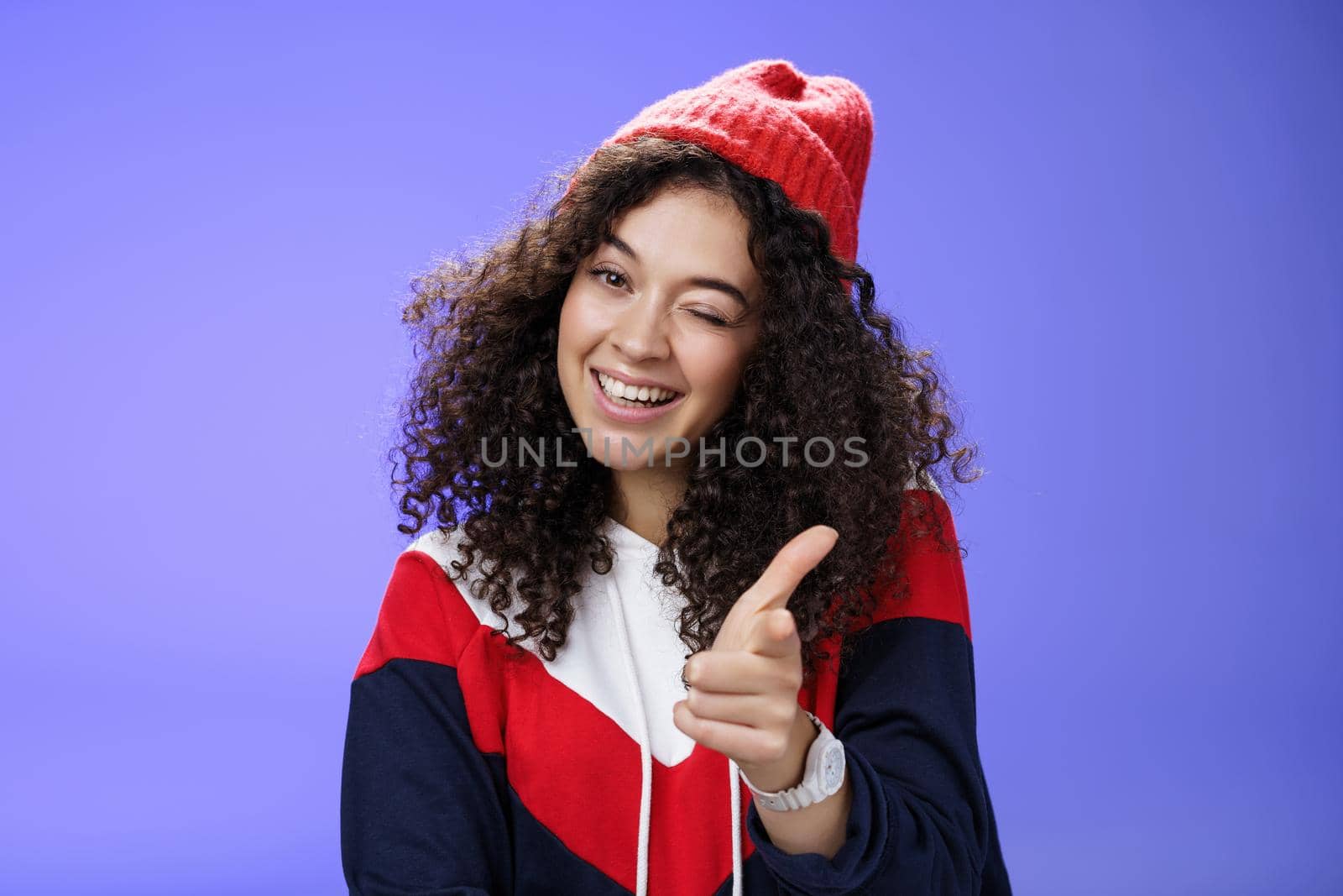 Friendly-looking sociable and stylish woman with curly hair in red beanie winking joyfully and pointing with finger pistol at camera as greeting mate being cool and confident over blue wall.