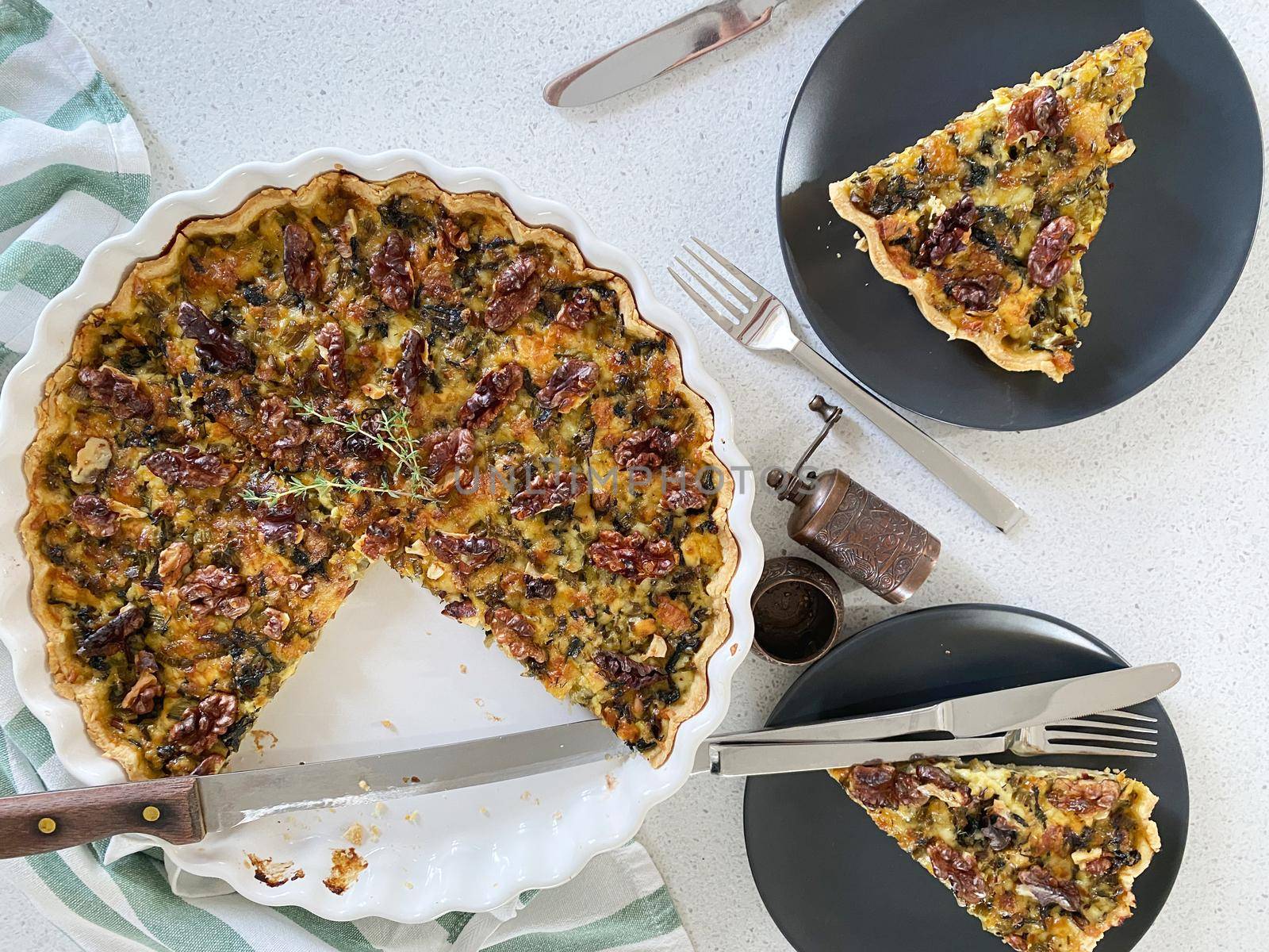 Quiche with ricotta and dried tomatoes in a large white baking dish. Two dark plates with portioned pieces of quiche, cutlery