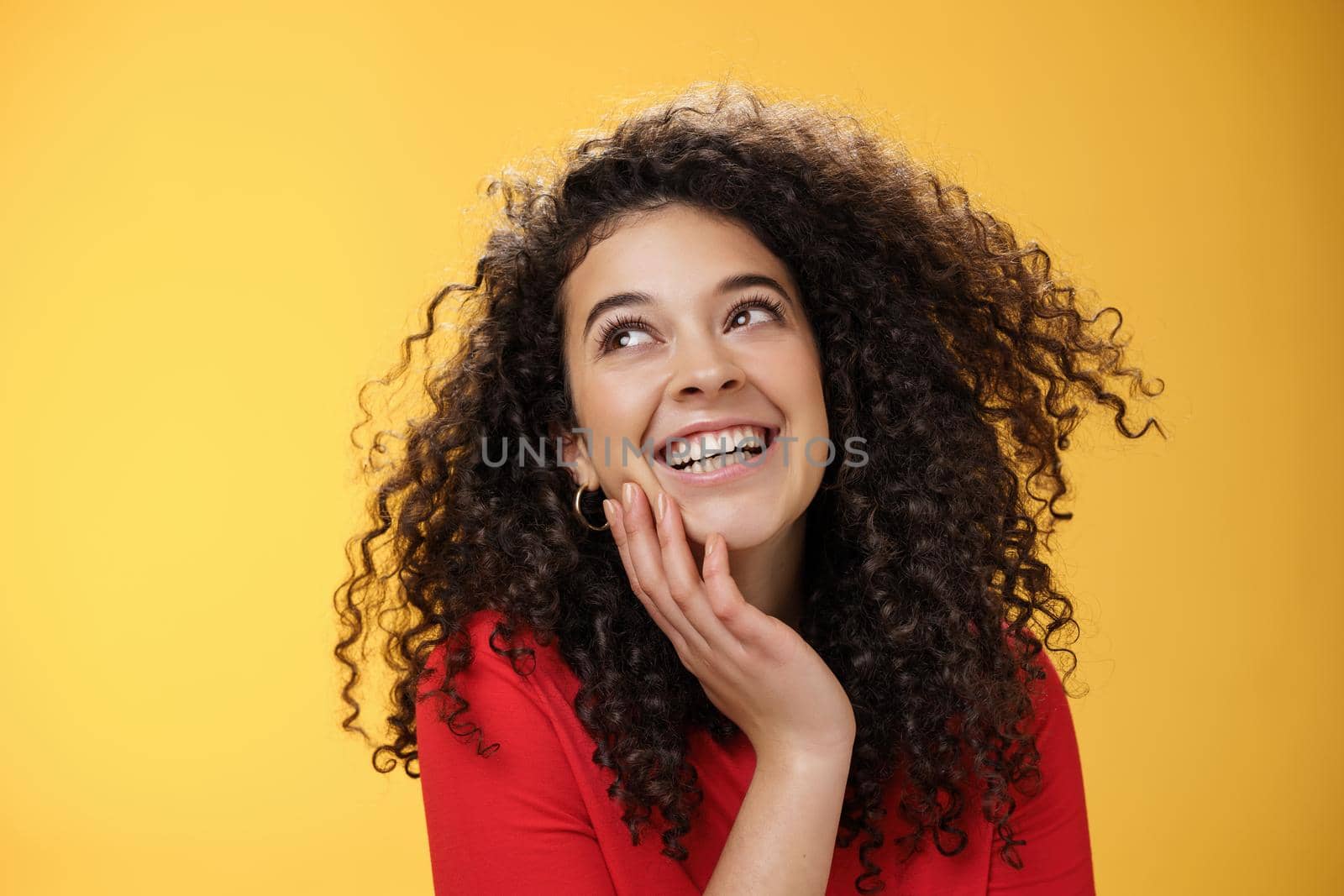 Happy positive pretty young woman with curly hair in red blouse laughing silly and carefree as gazing pleased at upper left corner, touching face, satisfied and delighted over yellow wall by Benzoix