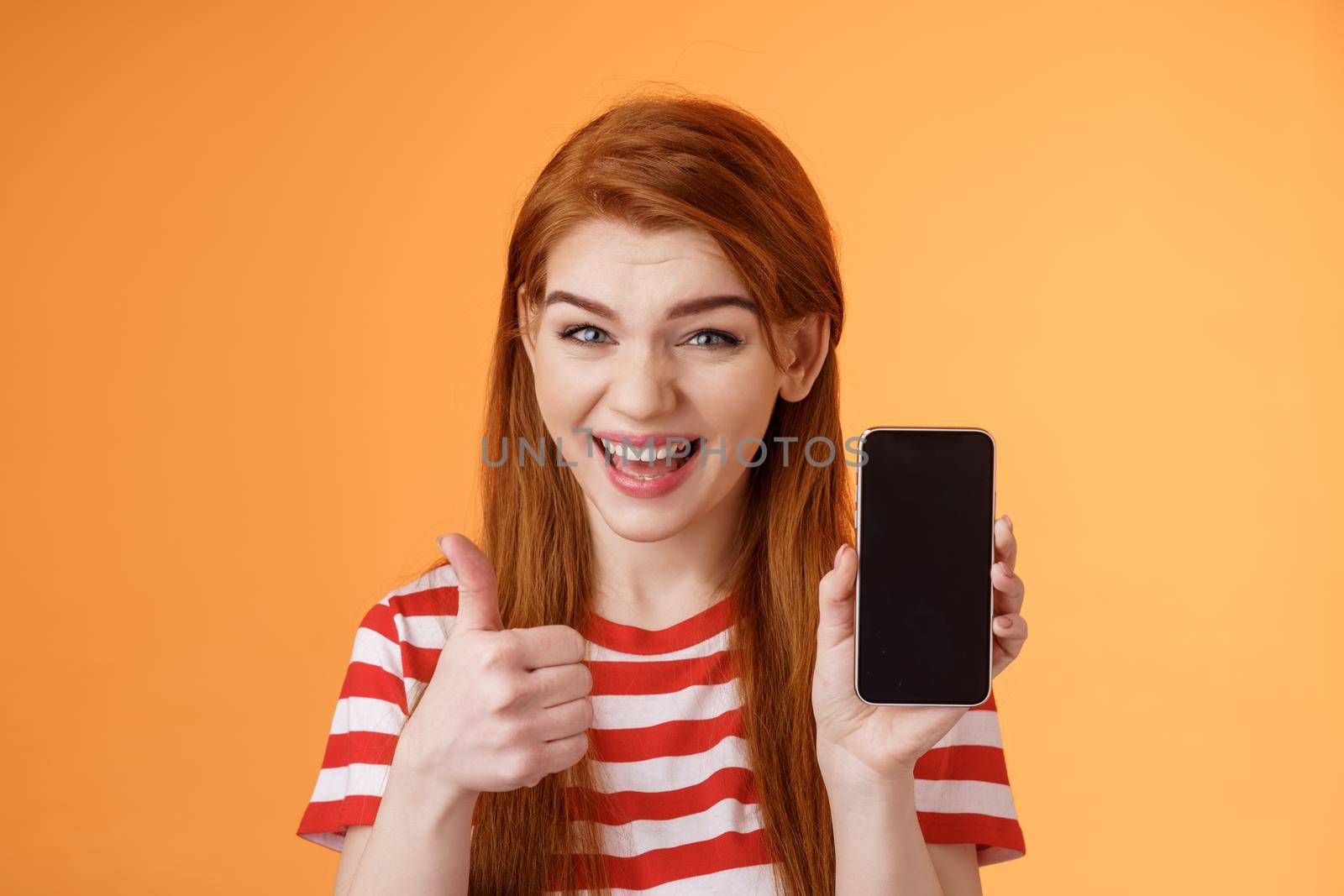 Cheerful assertive redhead woman recommend smartphone app, show phone display, thumb-up like approval sign, smiling broadly, suggest good online purchase, mobile advertisement. Copy space