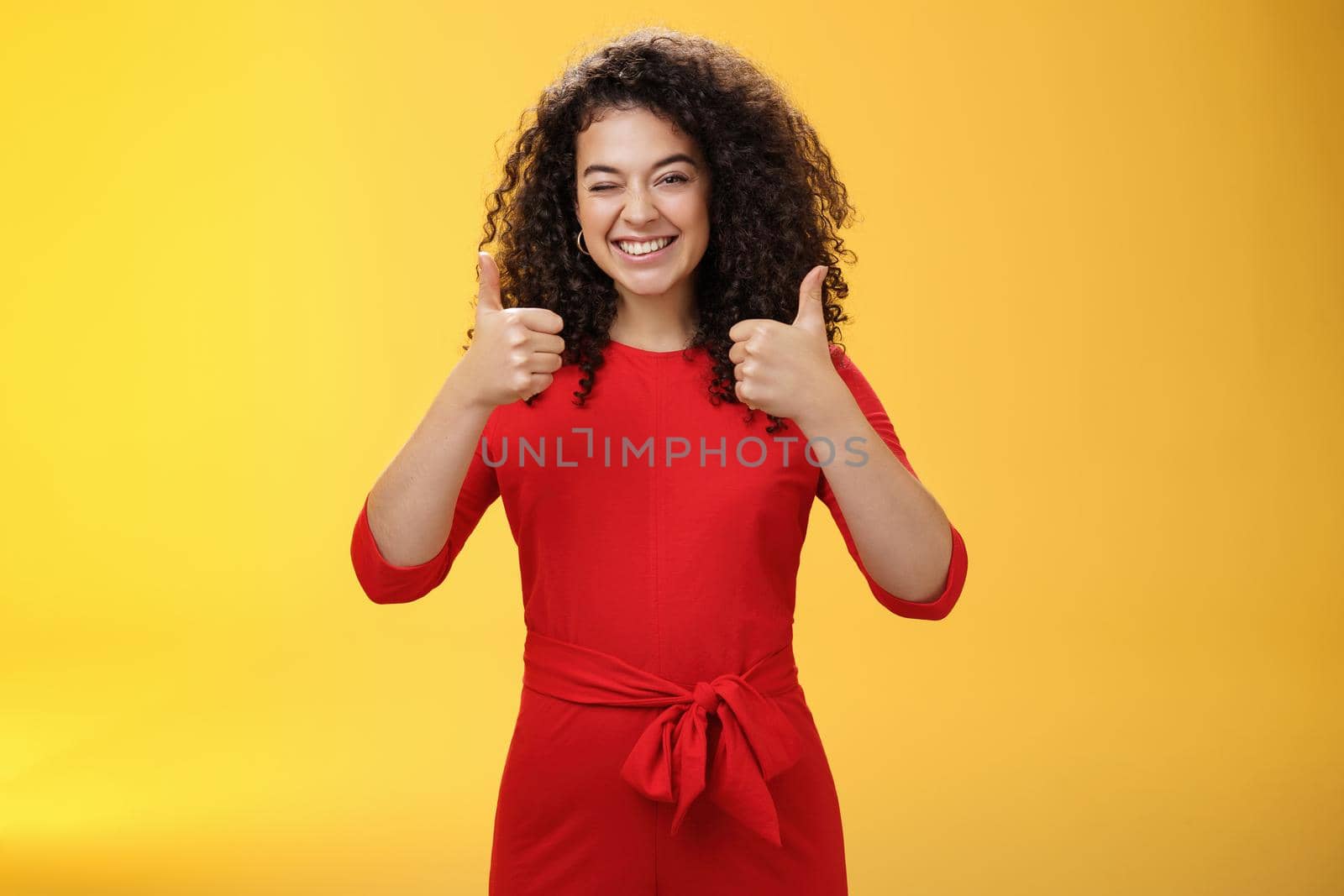 Creative and charismatic happy upbeat woman 25s with curly hair in red dress winking in approval and showing thumbs up with broad smile, satisfied giving positive reply over yellow wall by Benzoix