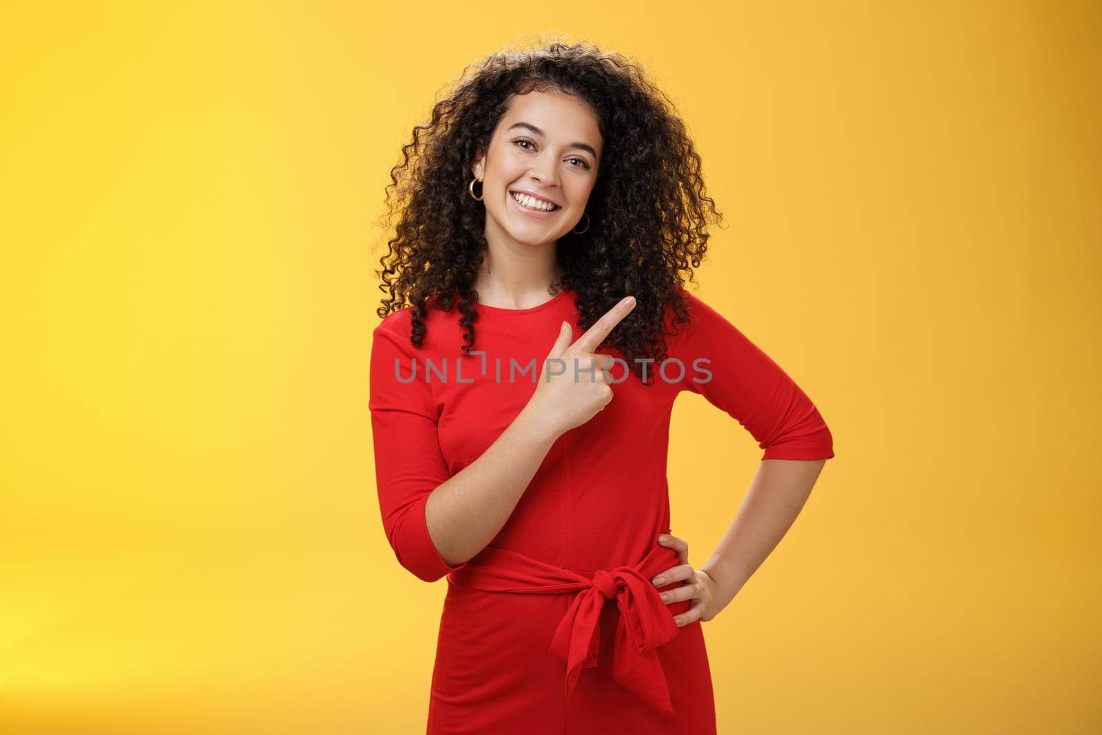 Girl suggesting try out new product. Friendly and pleasant happy young european brunette with curly hair tilting head cute, smiling broadly, pointing at upper right corner to direct at advertisement.