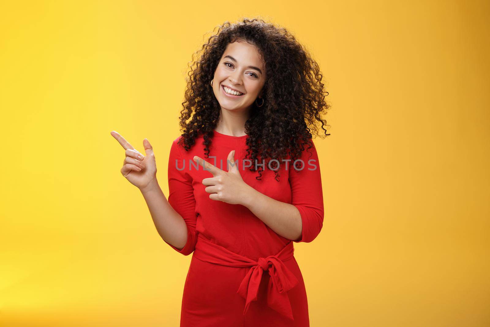 Friendly cute assertive female assistant in elegant red dress with curly hair tilting head flirty and smiling broadly, satisfied as pointing at upper left corner, recommending product over yellow wall.