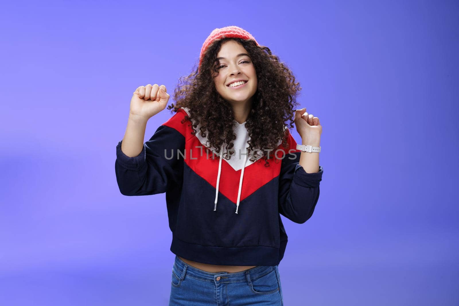Charming carefree young 20s girl with curly haircut in warm beanie dancing and having fun joyfully over blue wall raising hands relaxed, chill smiling broadly at camera liking perfect chilly weather.