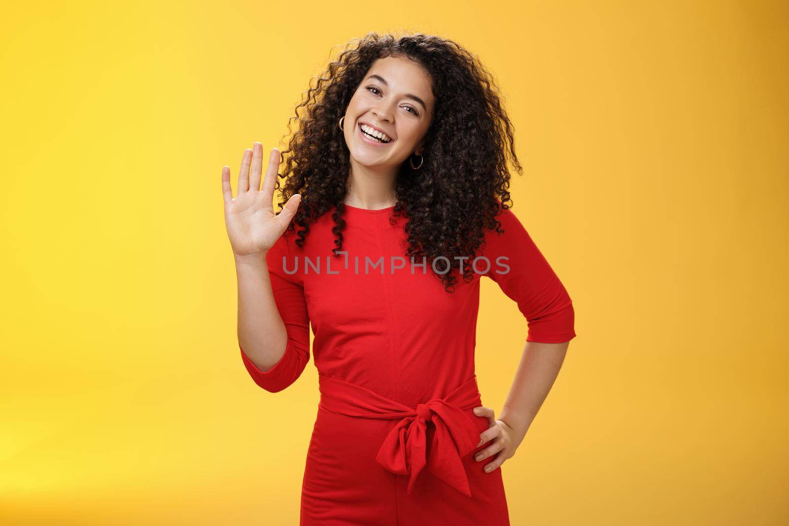 Hey my name is. Friendly-looking self-assured carefree cute 25s woman with curly hair waving with raised palm in hello or hi gesture smiling broadly greeting new coworkers over yellow background.