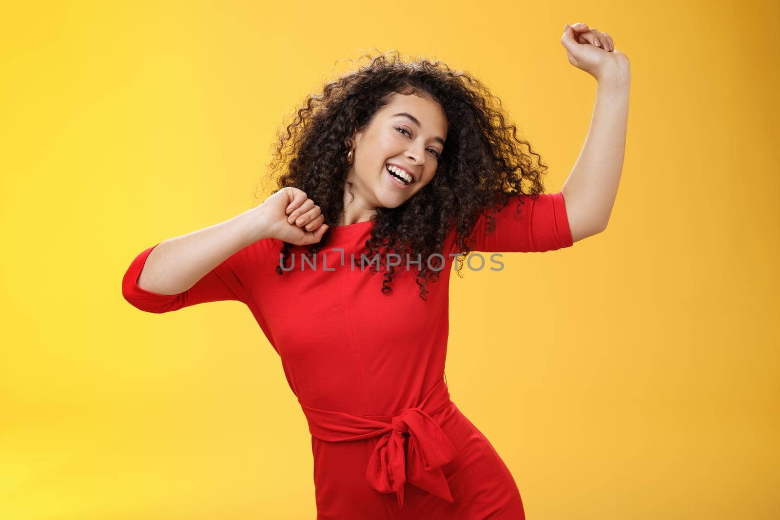 Carefree girl feeling uplifted and joyful dancing in red dress raising hands up happily tilting head and smiling broadly at camera as enjoying vacation, celebrating holidays over yellow background by Benzoix