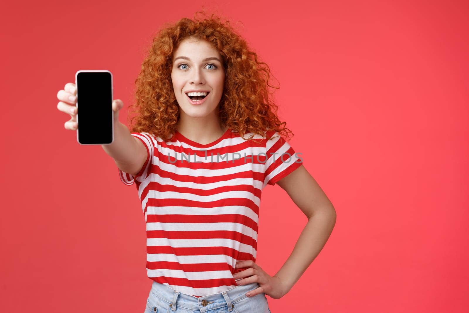 Cheerful attractive charismatic european redhead girl curly hairstyle show smartphone screen smiling happily promote app advice recommend good application social media page red background by Benzoix