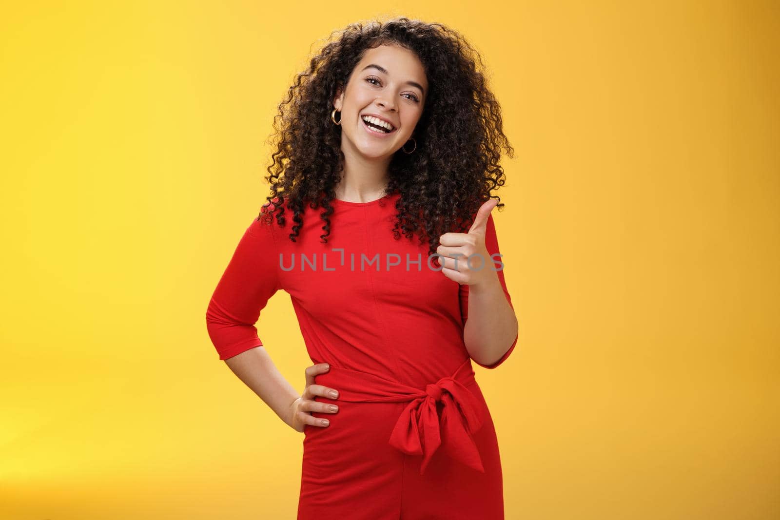 Portrait of happy optimistic young curly-haired woman in red dress laughing joyfully, showing thumbs up in approval and like gesture, delighted with awesome idea, accepting plan over yellow wall by Benzoix