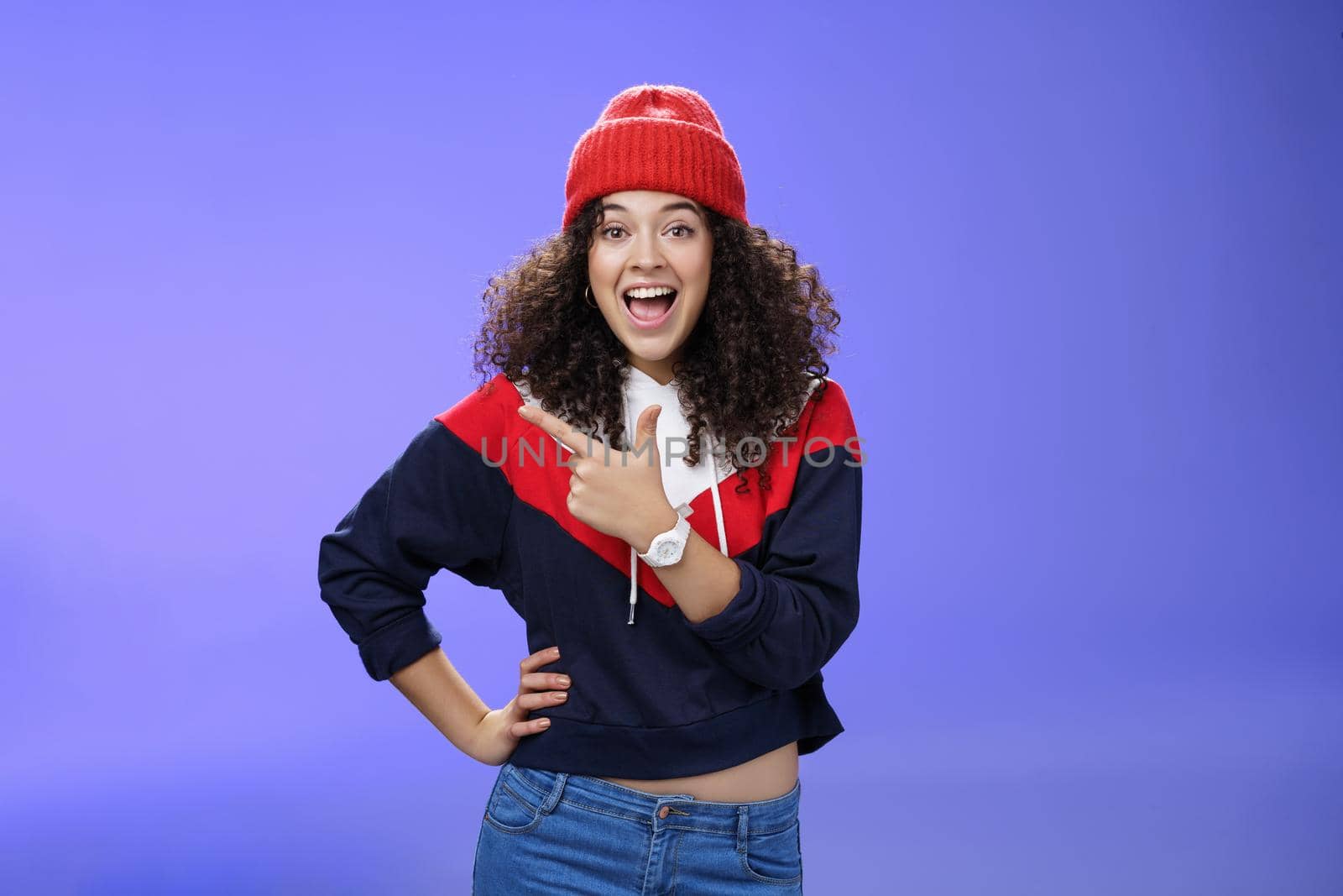 Portrait of enthusiastic and sociable european woman in warm hat and sweatshirt smiling delighted with amused grin as pointing at upper left corner impressed and astonished with awesome promotion.