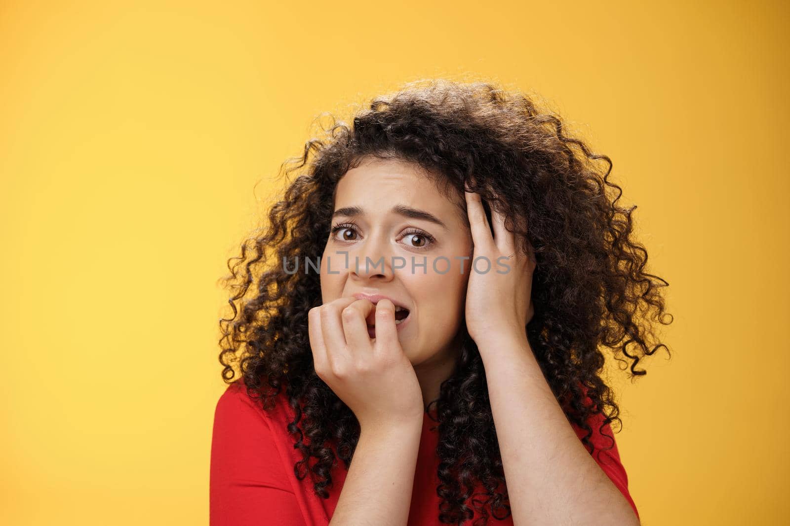 Scared woman overthinking frightened being in trouble biting fingers holding hand on head in despair, standing concerned, panicking and feeling uneasy as standing over yellow background. Emotions, facial expressions concept