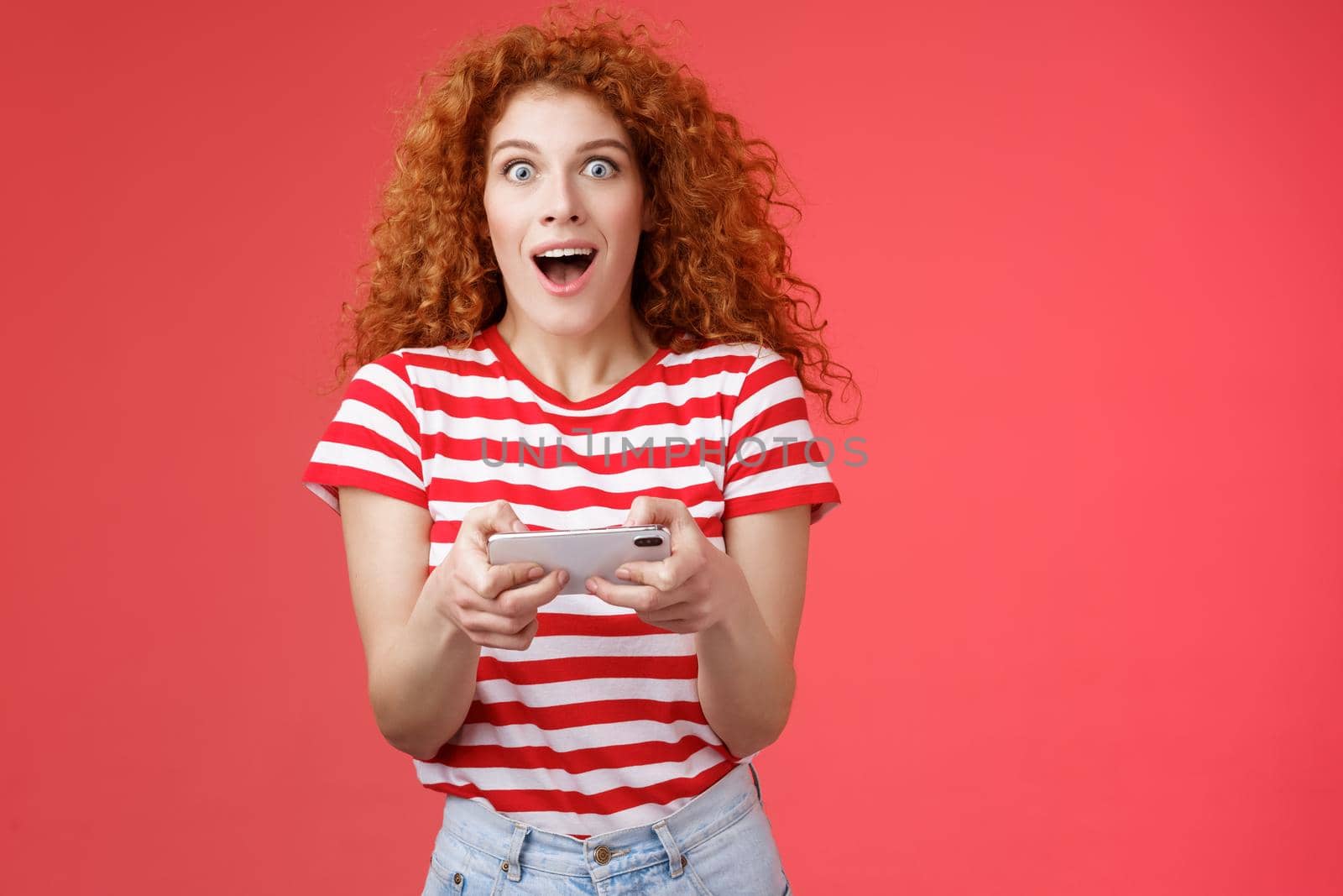 Amused impressed good-looking curly-haired redhead female student unexpectedly win smartphone game look camera astonished gasping open mouth stare camera hold phone horizontal playing app by Benzoix
