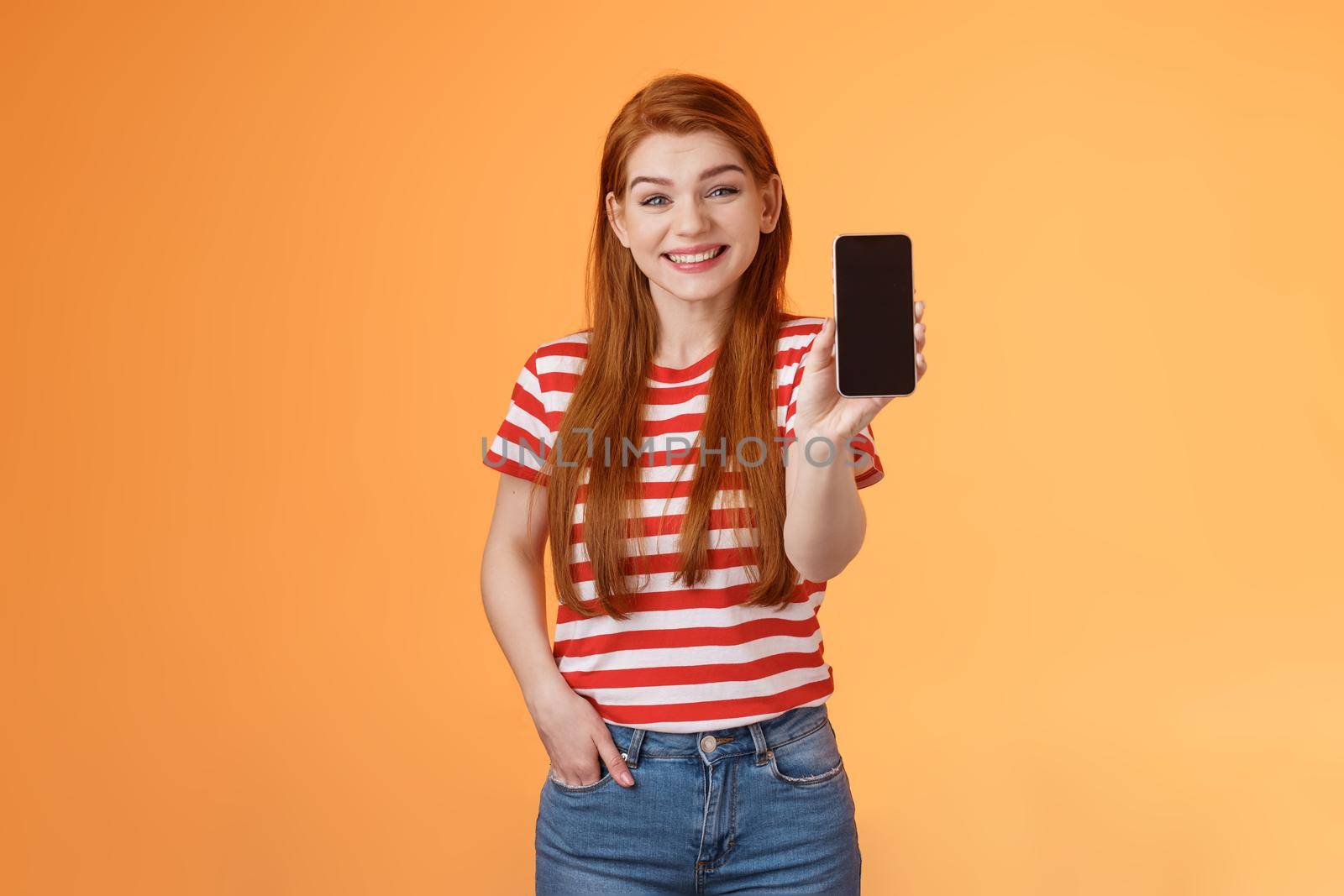 Cheerful good-looking 20s redhead woman introduce social media feature product hold smartphone, show camera cellphone display, promote device application, smiling broadly, orange background.