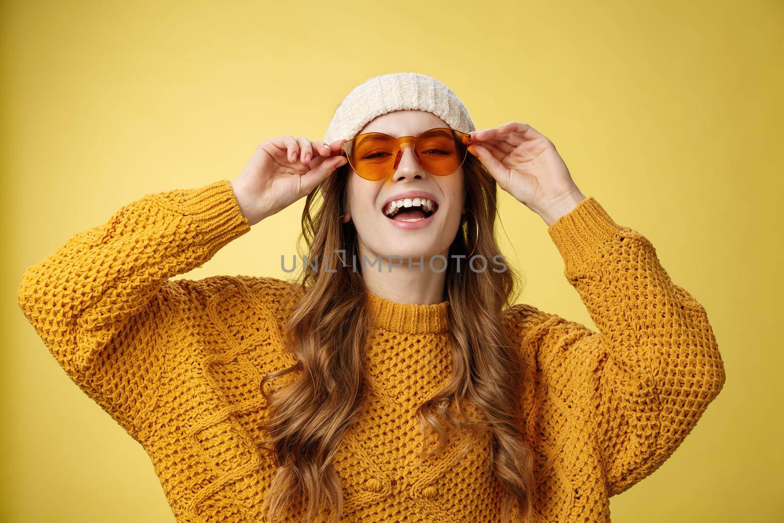 Joyful stylish caucasian girlfriend enjoying sunlight winter ski resort wearing sunglasses hat warm sweater having fun smiling touching eyewear, expressing playful happy mood yellow background.