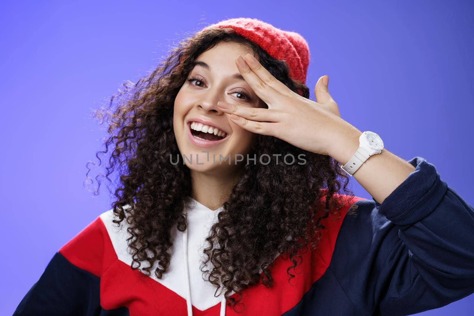 Close-up shot of charismatic happy carefree woman in winter red cute beanie and sweatshirt holding fingers near eye and peeking with broad smile at camera having fun, playing over blue background.