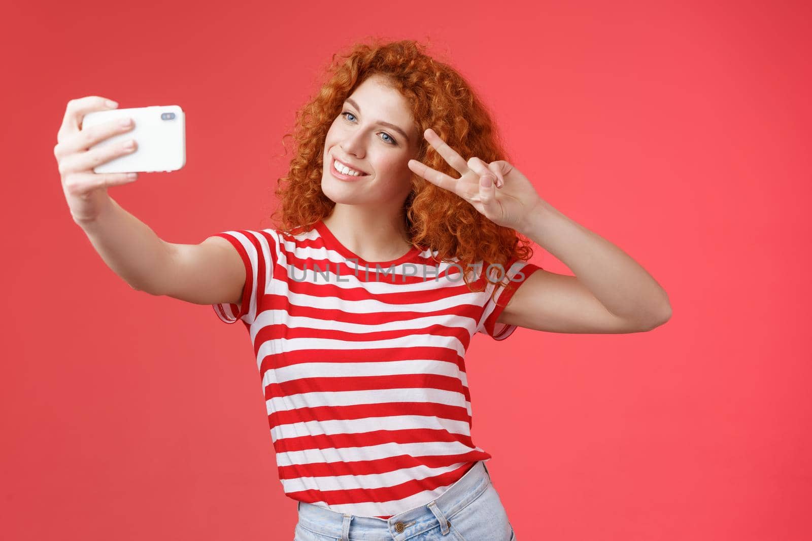Stylish carefree good-looking redhead curly woman enjoy summer holidays show victory peace gesture eye tilt head cute record video hold smartphone taking selfie red background. Copy space