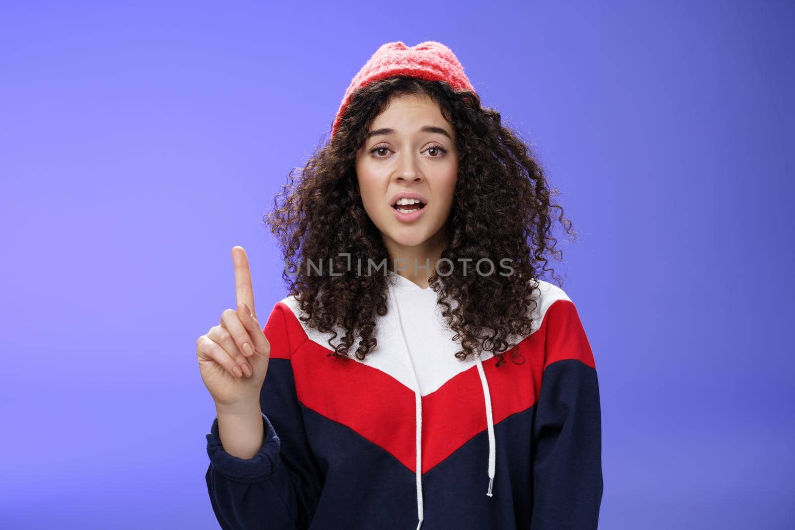 Rule number one do not mess with me babe. Portrait of swag and cool stylish young curly-haired woman in winter beanie showing index finger in prohibition or warning gesture over blue background by Benzoix