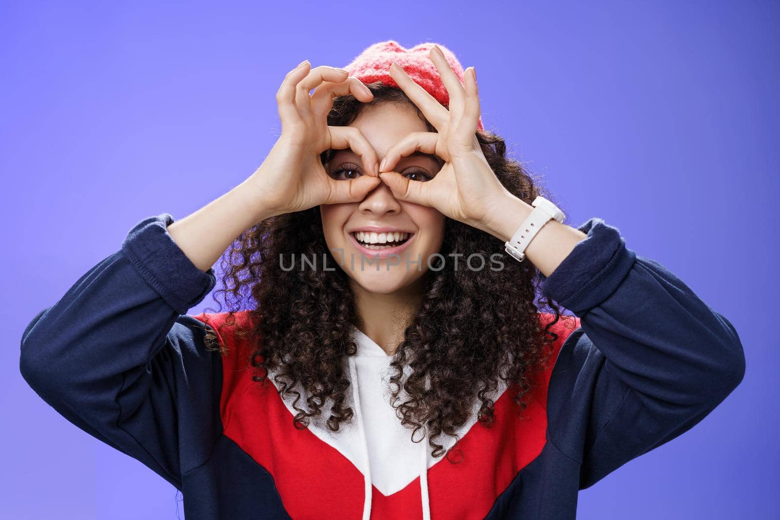 Girl see us as imitating peek with binocular making circles over eyes and looking through it joyful and excited, having fun fooling around posing happy and cute over blue background in outdoor outfit.