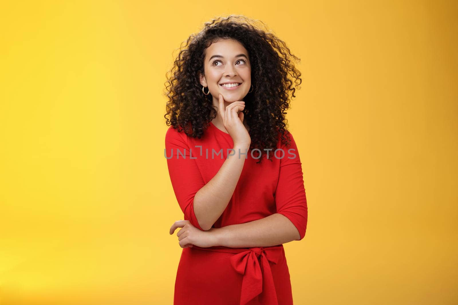 Girl having interesting idea, smiling as feeling confident plan might work. Charming happy woman with curly hair in red dress gazing at upper left corner thoughtful, thinking over yellow wall by Benzoix