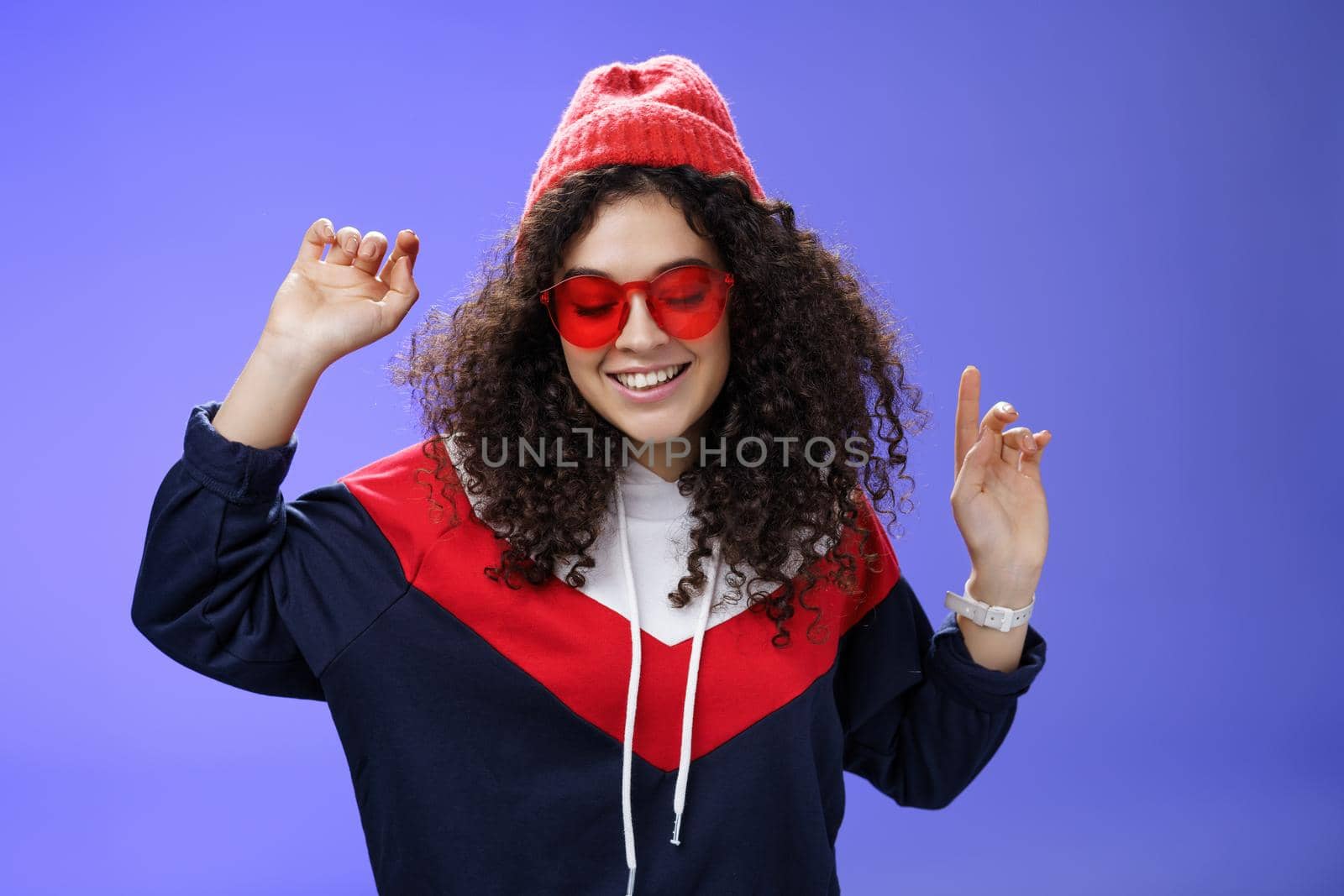 Girl feeling awesome having fun and enoying cool party, dancing with raised hands looking down smiling broadly wearing sunglasses and stylish beanie posing against blue background by Benzoix