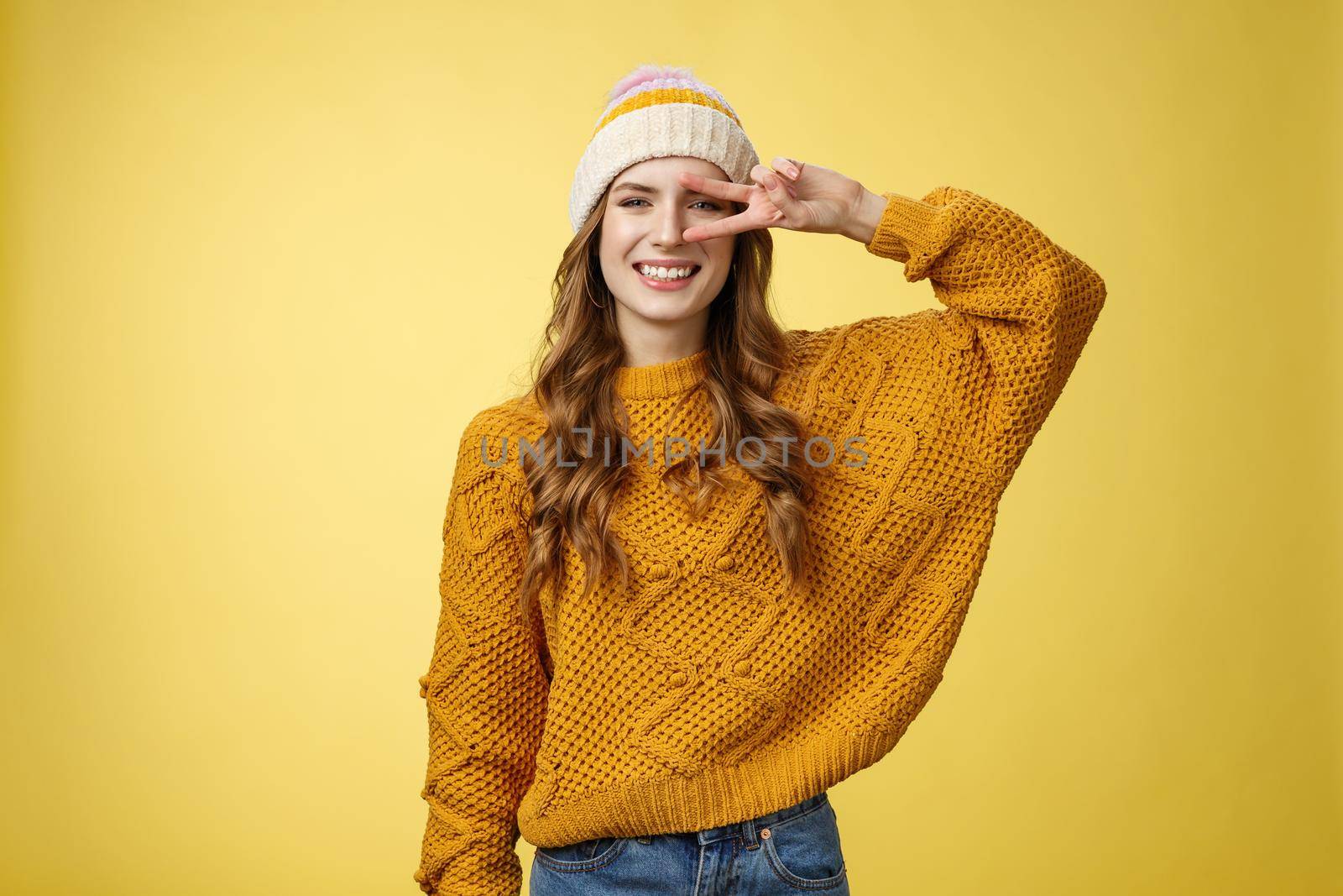 Positive peaceful kind charming young woman show peace victory gesture near eye smiling broadly have good mood perfect day wishing you good luck, standing cheerful grinning yellow background.