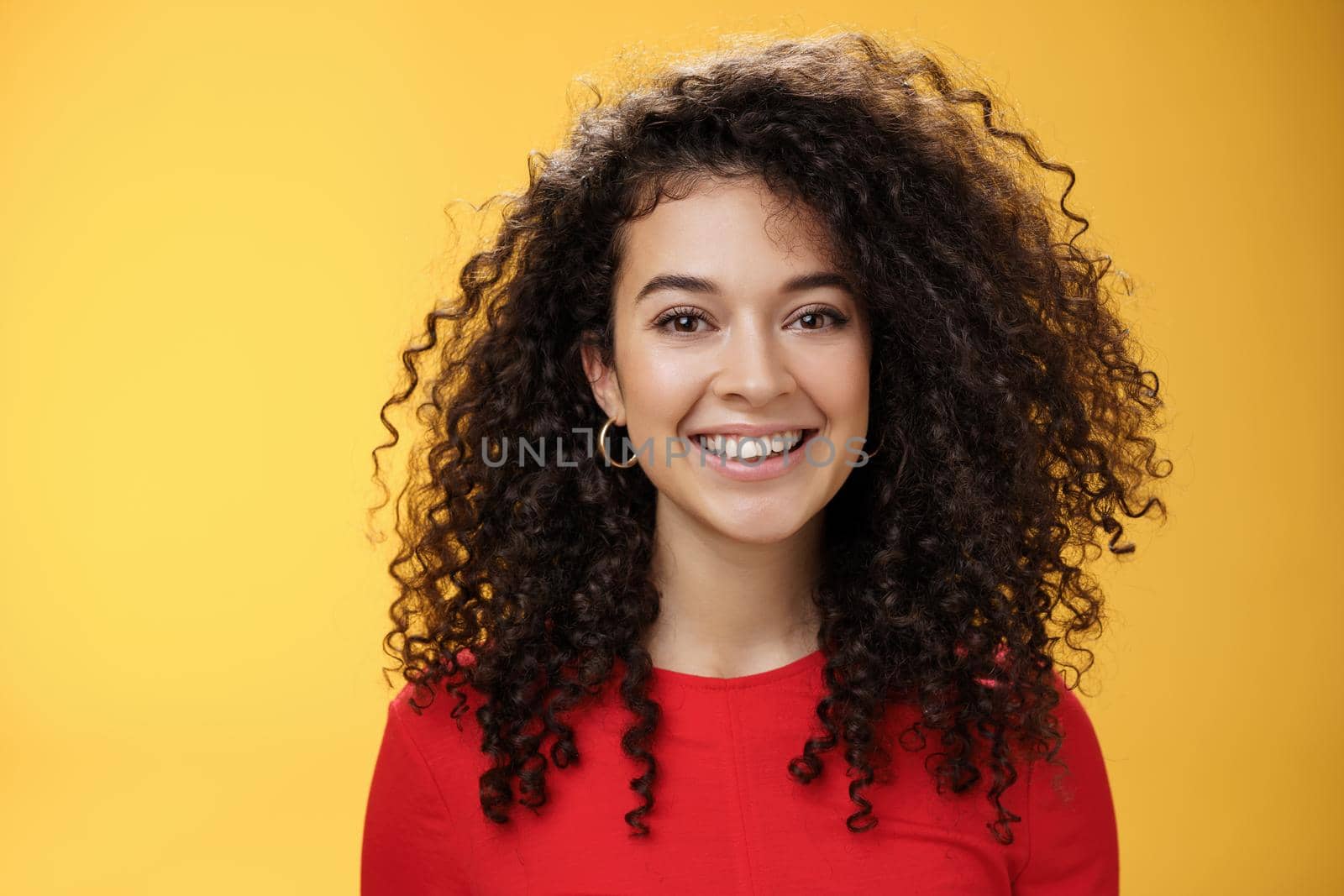 Close-up shot of pretty caucasian girl with curly hair in red dress and earrings smiling joyfully with pleased hopeful expression gazing at camera carefree, posing over yellow background by Benzoix