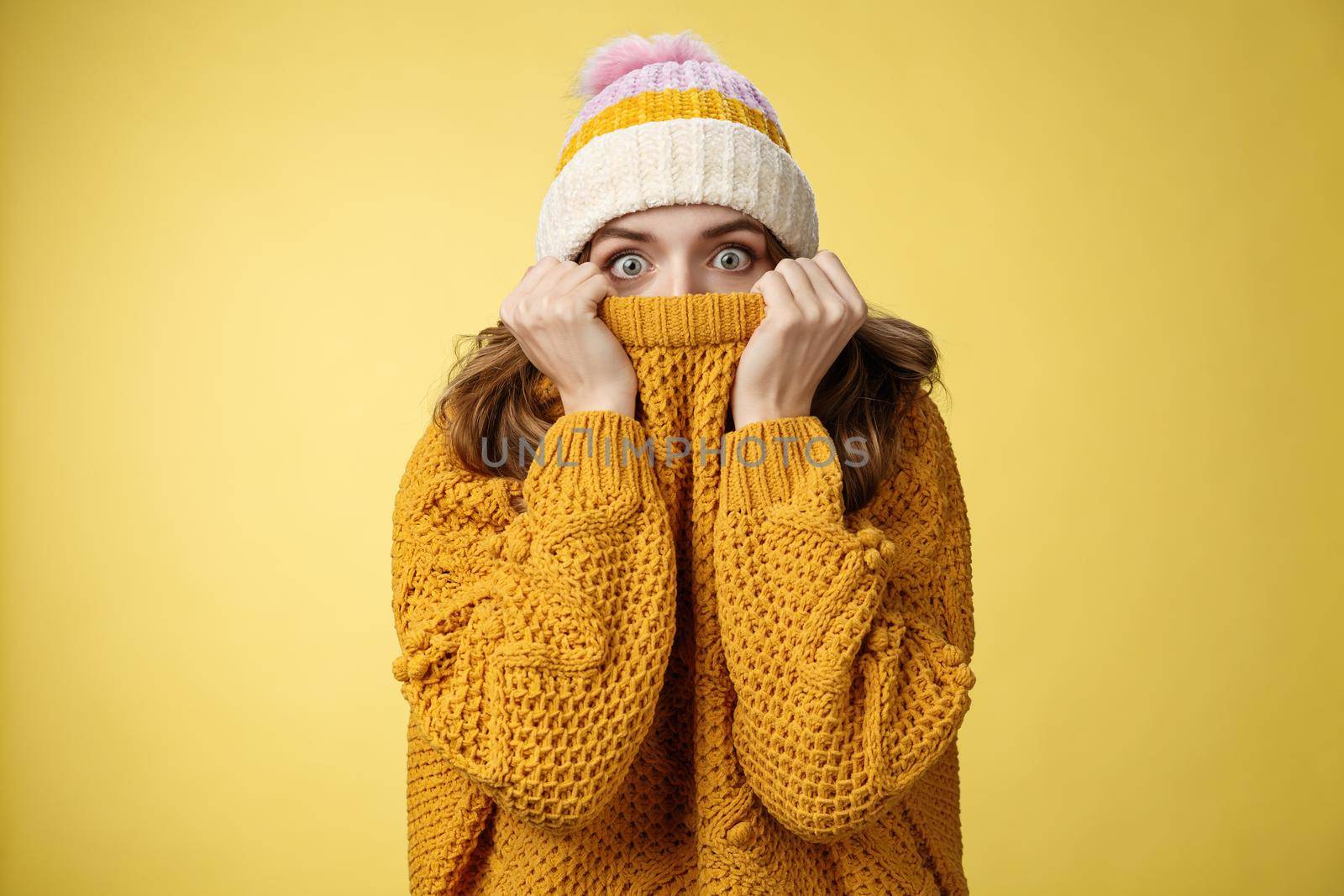 Portrait scared insecure timid cute girl hiding face pull sweater nose widen eyes afraid stunned standing stupor yellow background wearing winter hat, frightened terrified cold weather.