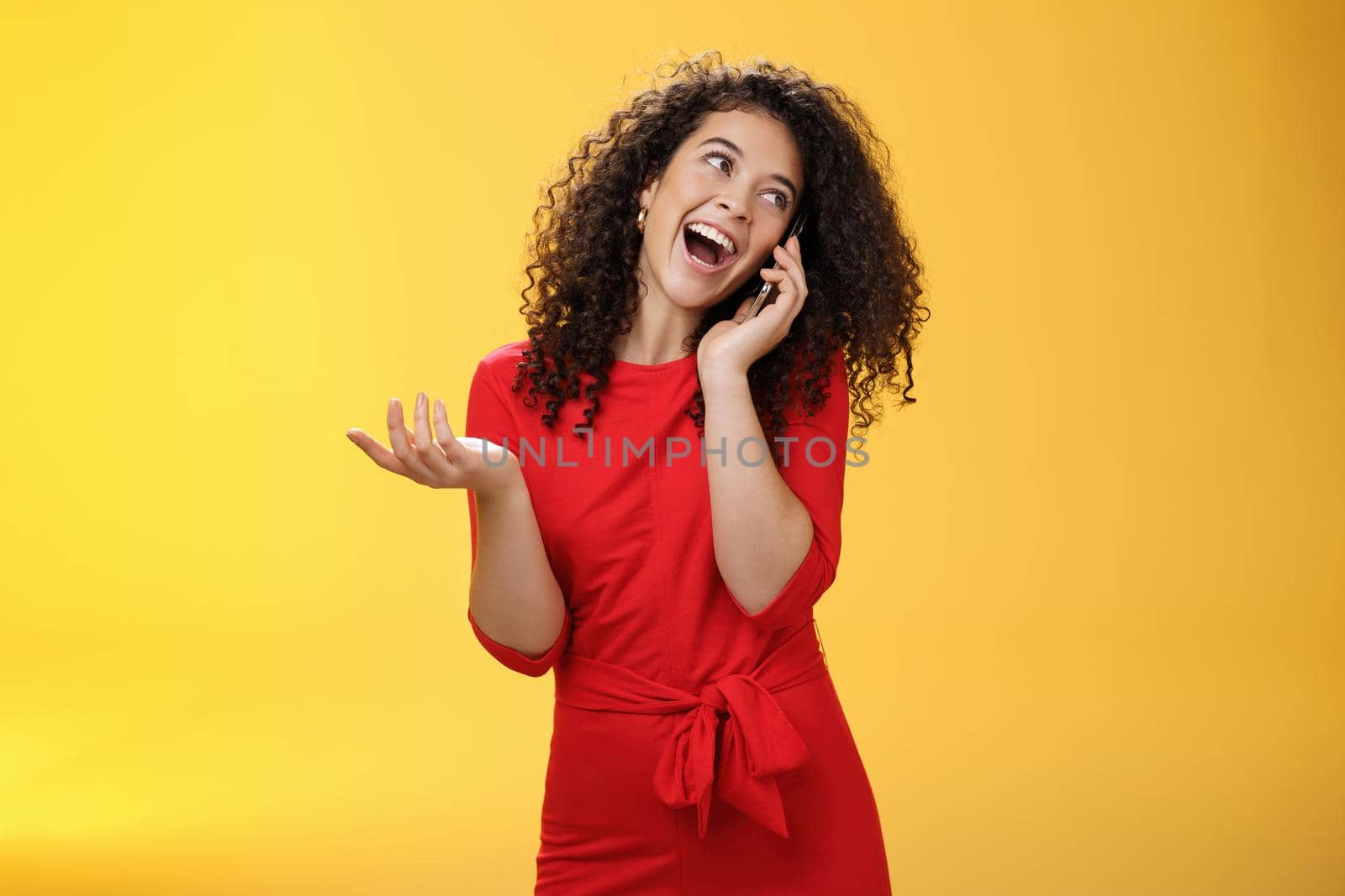 Talkative glamour silly girl with curly hair having fun feeling carefree and happy taling on mobile phone turning away as laughing joyfully gesturing with hand holding smartphone pressed to ear by Benzoix