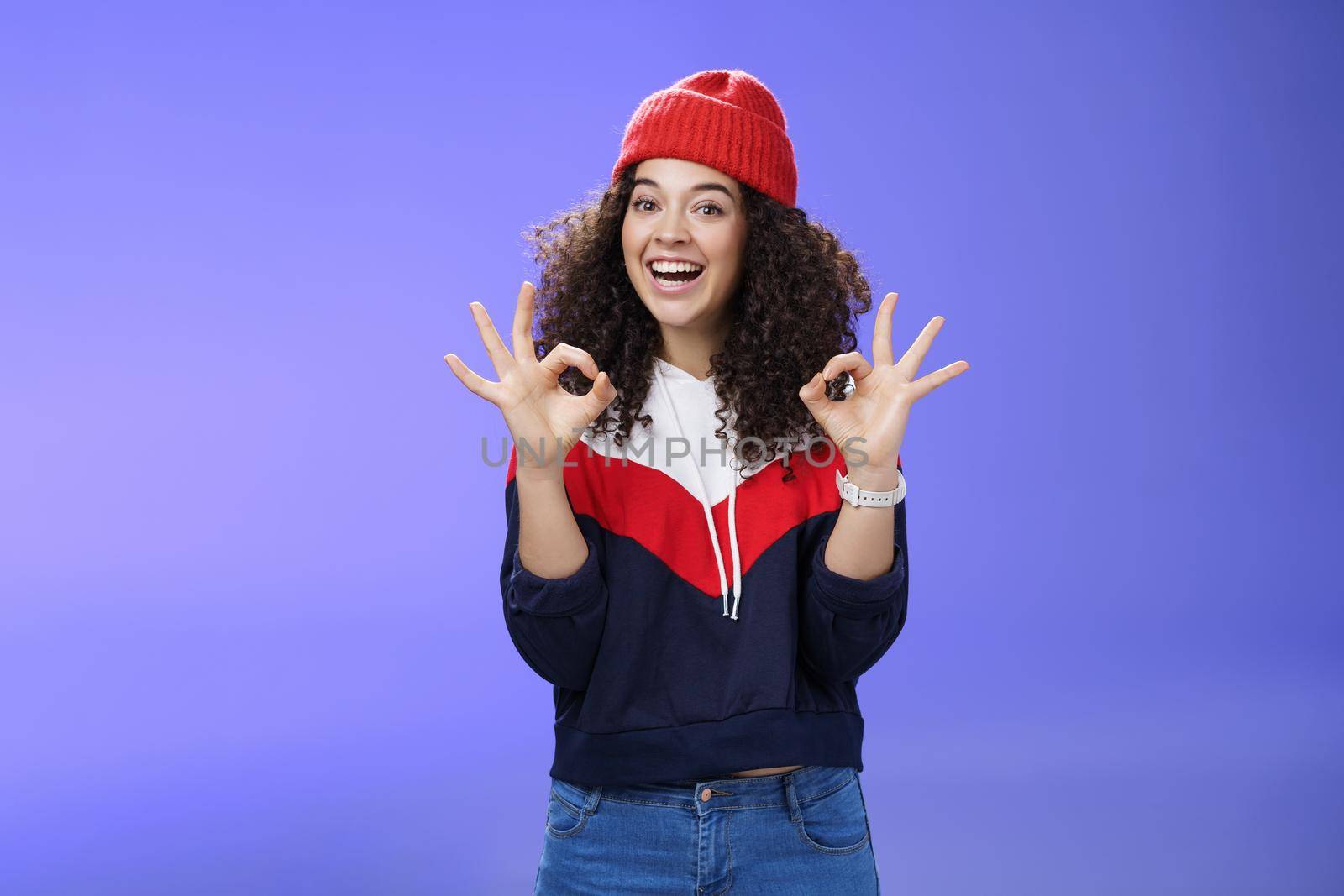 Got under control. Portrait of happy charming smiling curly-haired female in warm winter hat and sweatshirt smiling broadly and showing okay or excellent gesture as approving, liking cool movements.