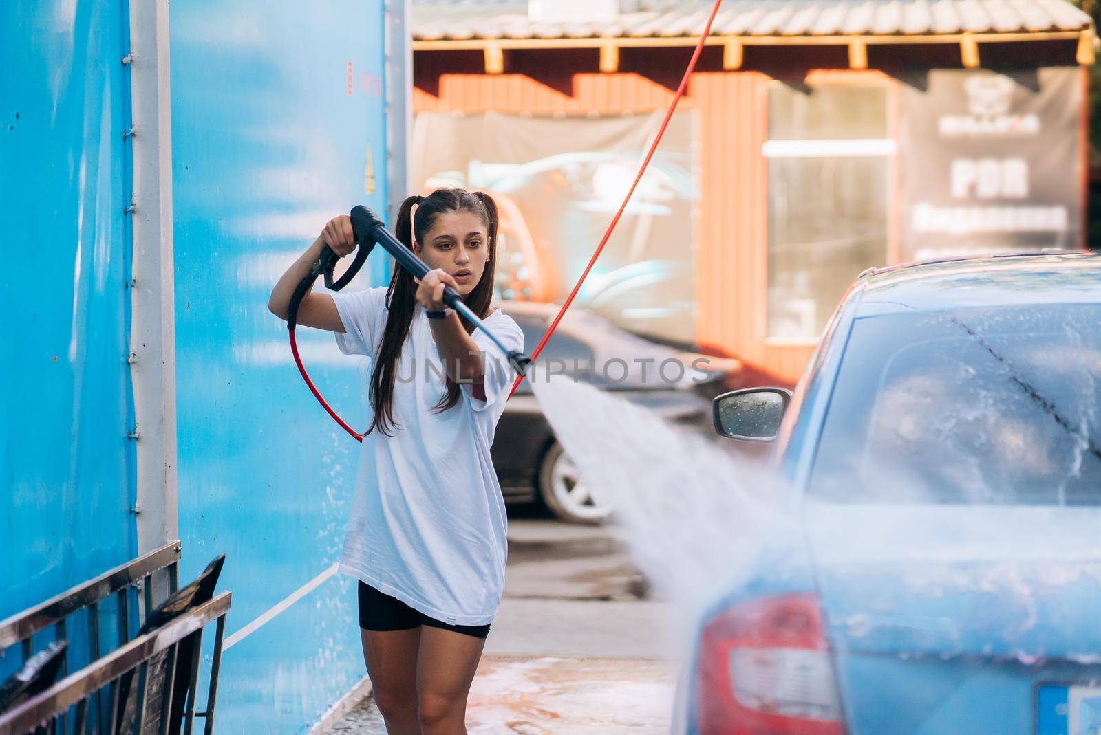 Brunette with two pigtails from a high-pressure hose washes the car at a car wash