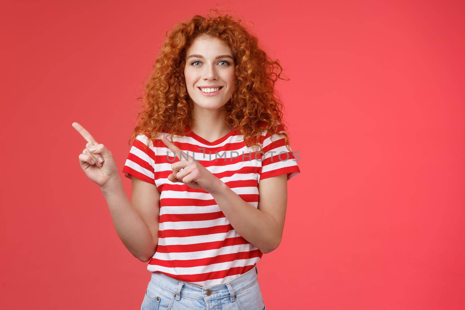 Joyful friendly attractive helpful cute redhead european female advice cool store promo pointing upper left corner blogger showing around perfect place spend summer holidays, red background.
