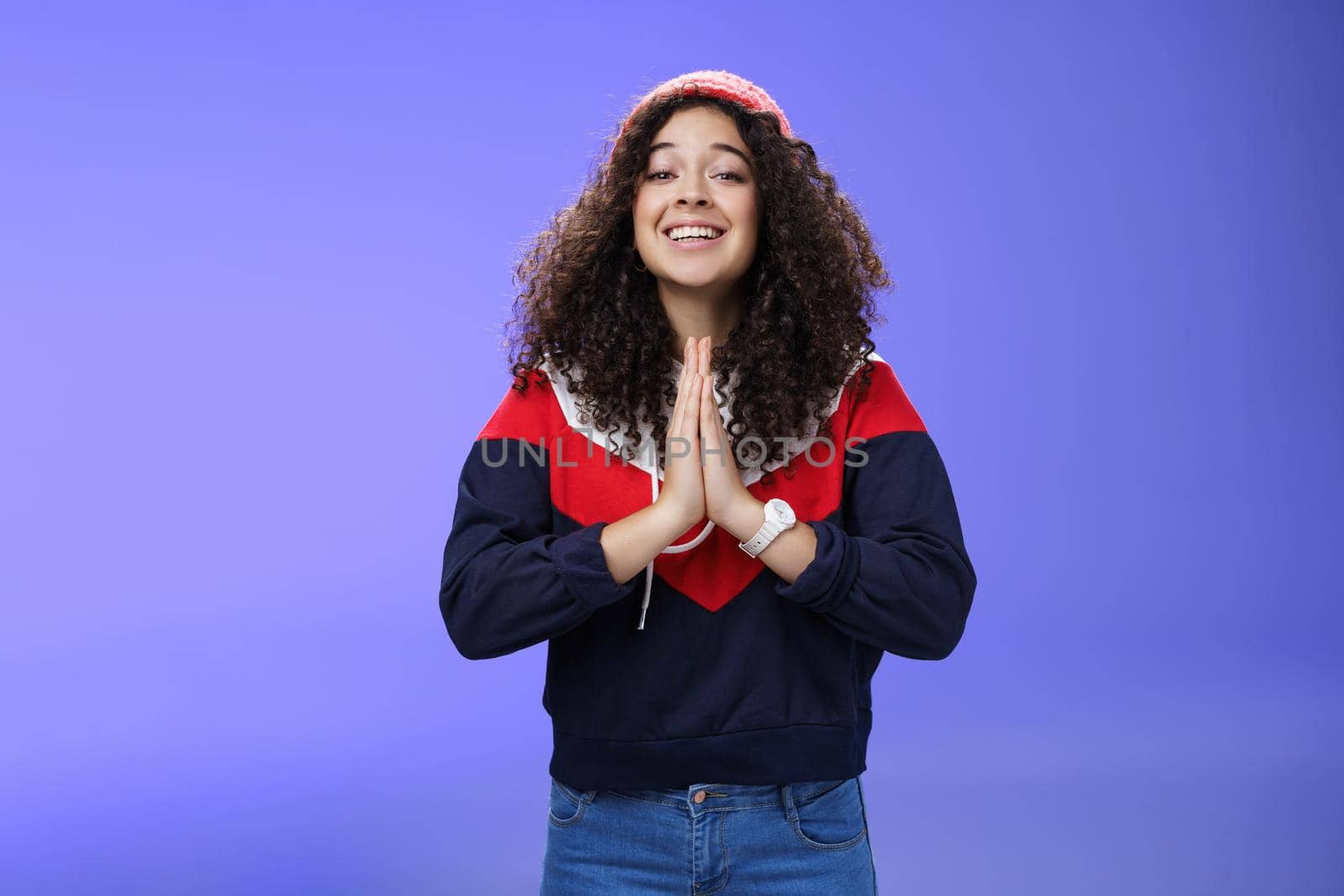 Charming excited and friendly-looking cute female friend with curly hairstyle in outdoor hat and sweatshirt holding hands in pray and smiling with angel expression as hoping friend help over blue wall.