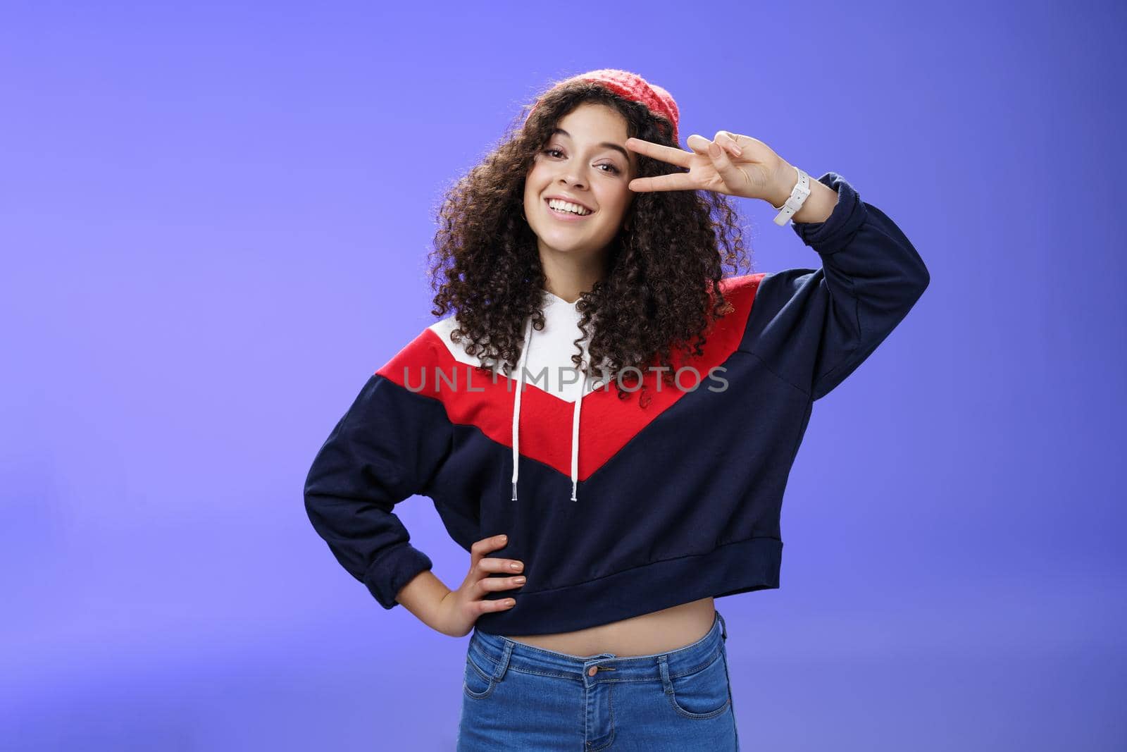 Attractive feminine woman with curly hairstyle in hat and sweatshirt showing peace or victory sign around eye and smiling carefree having fun playing in yard with snow over blue background by Benzoix