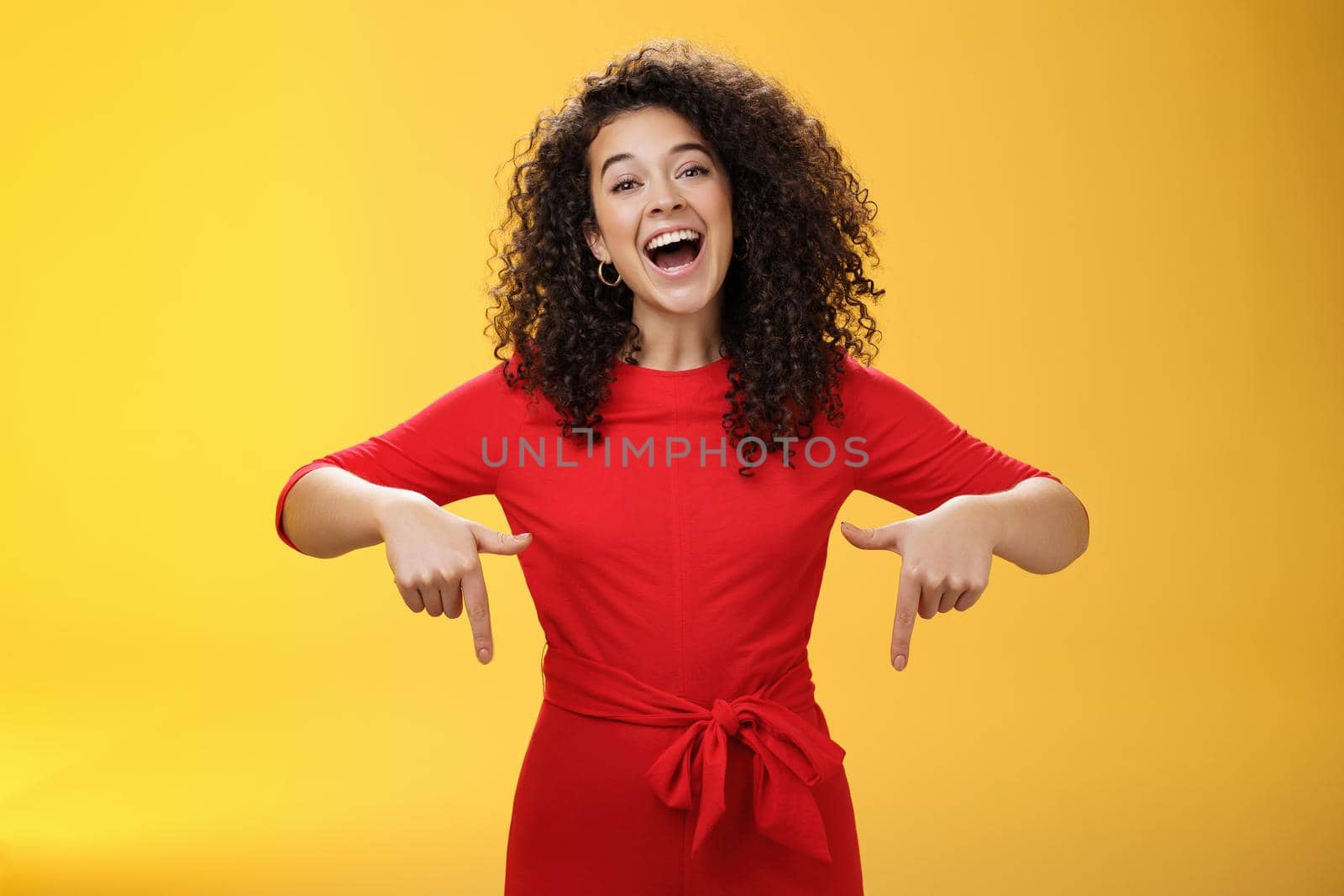 Look what I got here. Charismatic carefree happy charming woman with curly hair in red dress laughing with broad smile pointing down as showing awesome copy space to customers over yellow wall by Benzoix