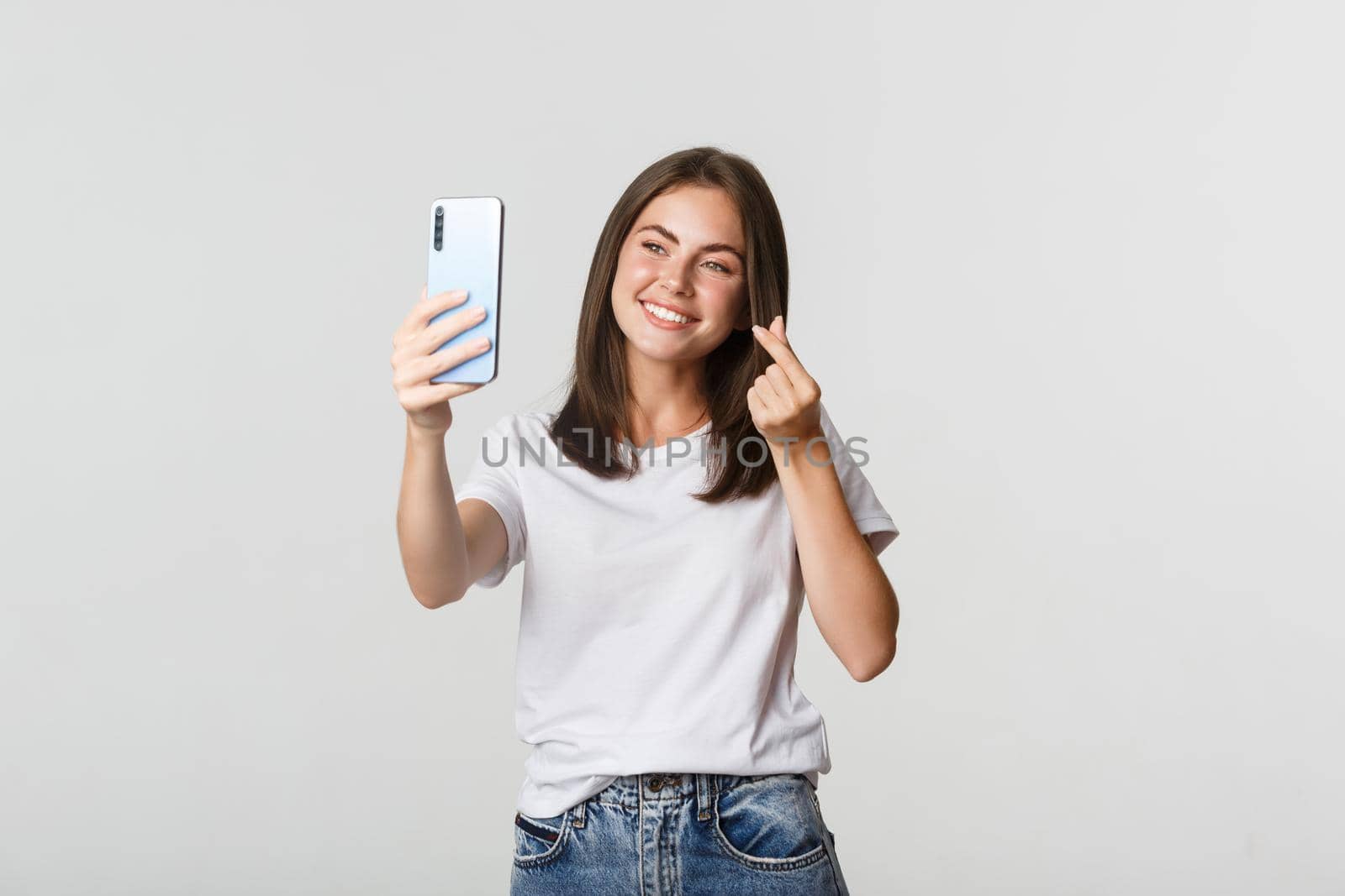 Happy beautiful young woman showing heart gesture and taking selfie on smartphone and smiling.