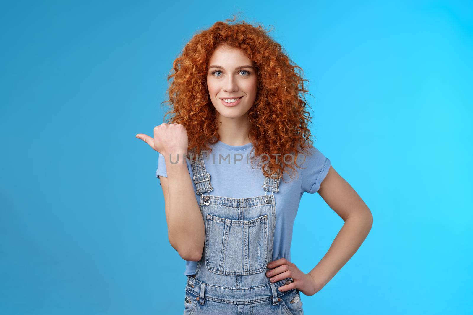 Cheerful curious lively redhead good-looking curly-haired girl wearing summer overalls t-shirt pointing left thumb look camera intrigued excited explore new stores friends walking shopping mall.