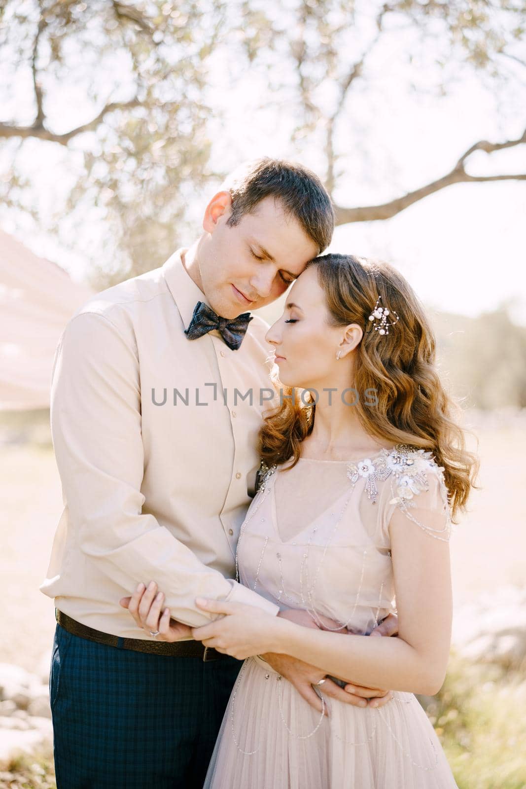 Groom hugs bride while standing under the tree. Portrait by Nadtochiy