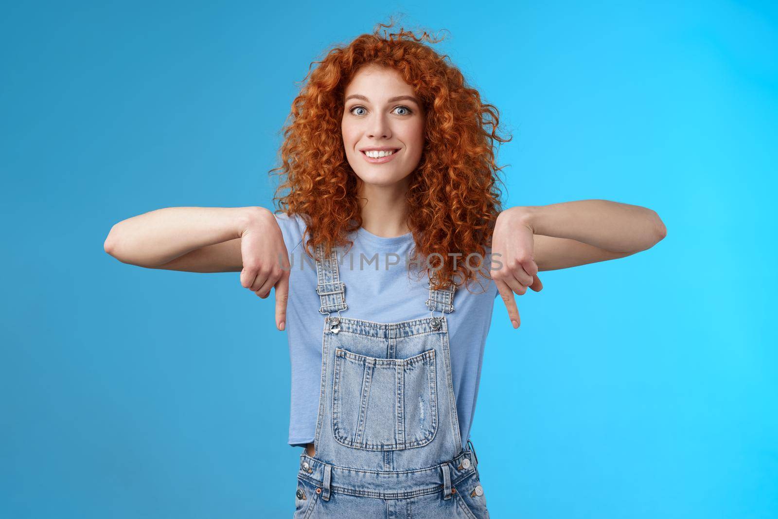 Excited happy smiling caring redhead curly-haired girlfriend thrilled awaiting summer holiday trip pointing down index finger upbeat mood show favorite store buy best prices, blue background by Benzoix