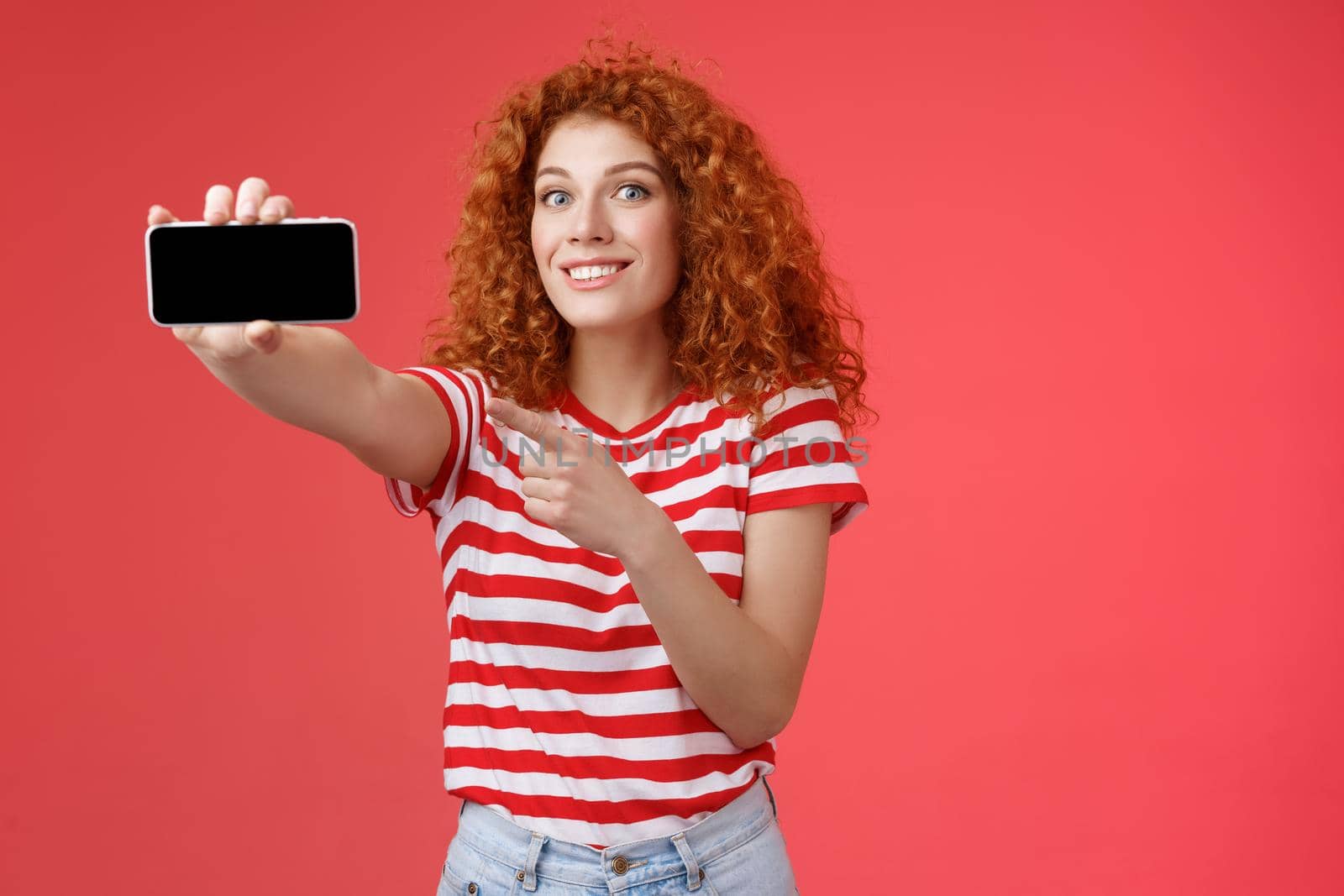 Excited happy good-looking redhead curly female show horizontal smartphone screen pointing display gadget smiling pleased proud beat friends score game standing red background by Benzoix