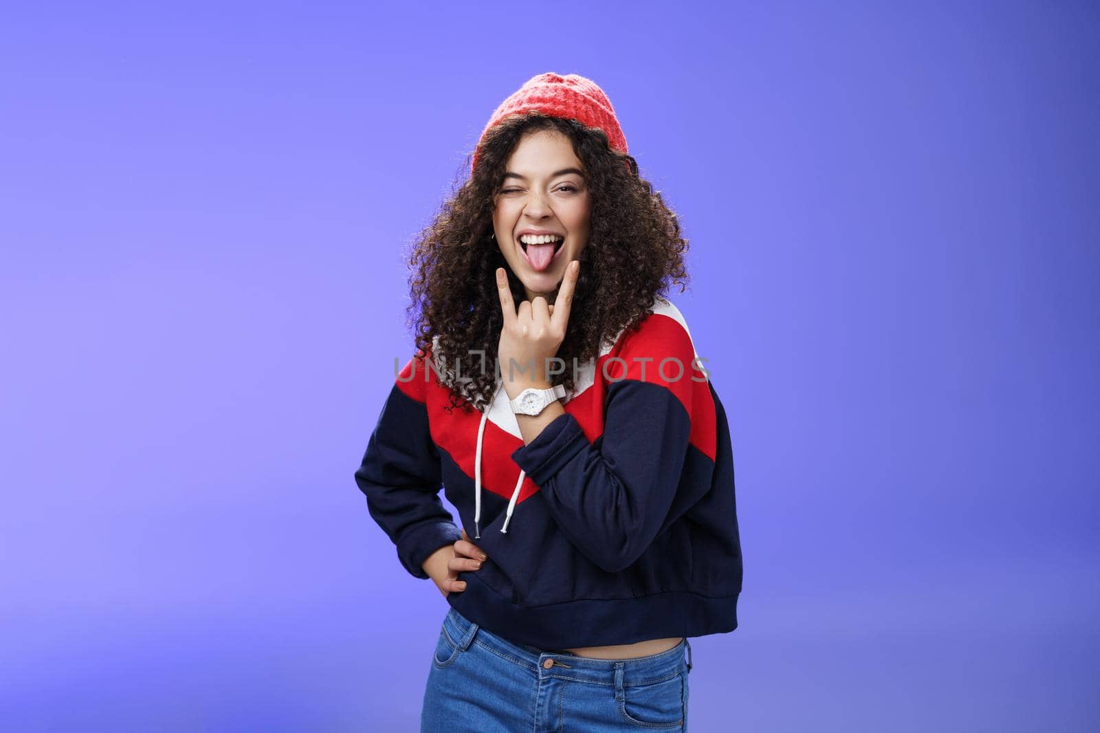Stylish and cool energized swag girl with curly hairstyle in warm beanie showing rock-n-roll gesture near mouth and sticking out tongue smiling having fun, partying and enjoying life over blue wall.