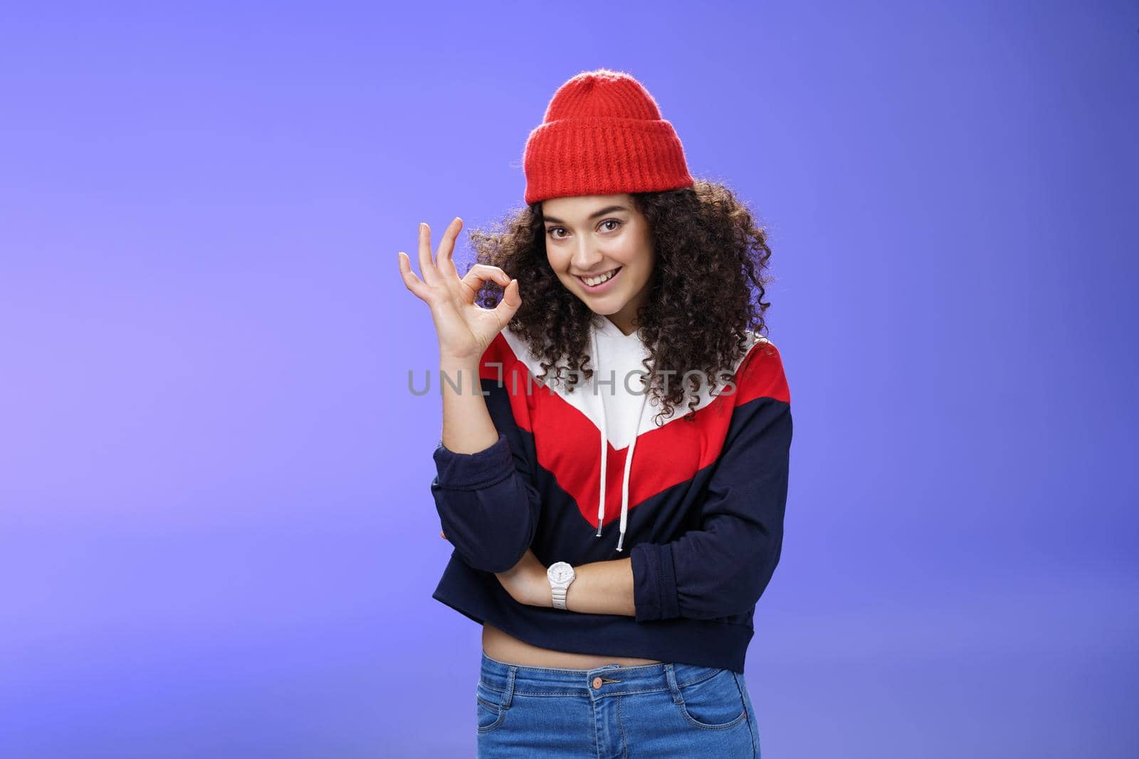 Portrait of pleased good-looking assertive woman with confident look and curly hair wearing warm winter hat showing okay gesture and smiling assuring, liking and approving awesome outfit of friend by Benzoix