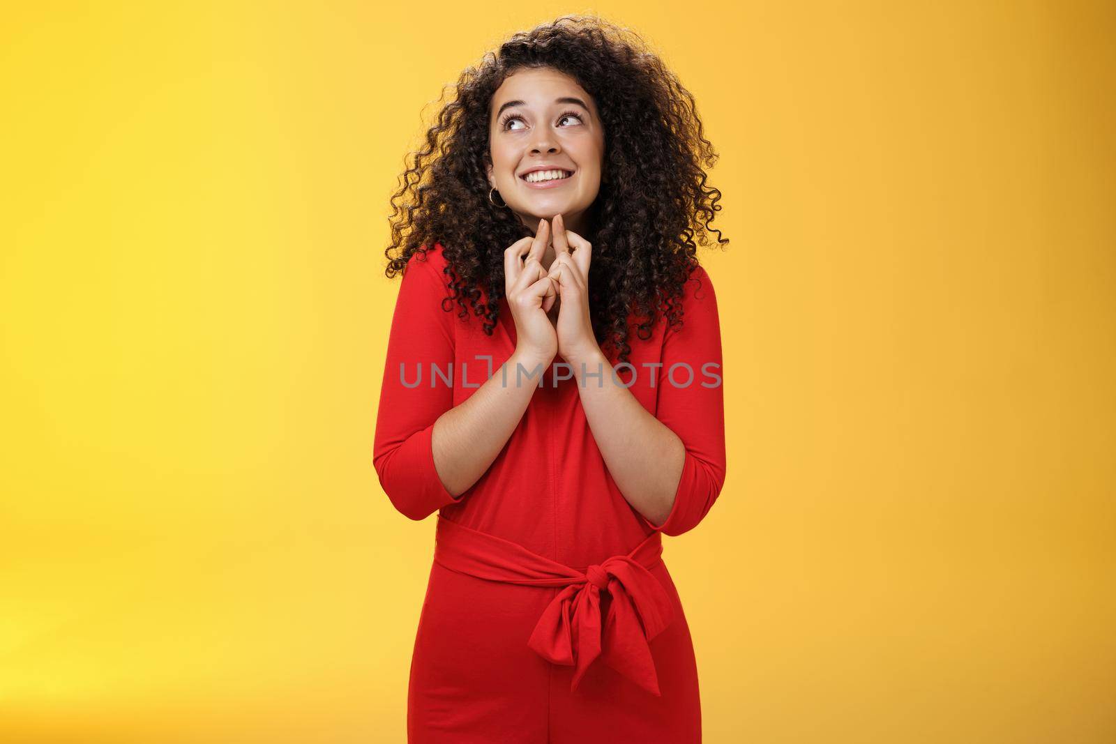 Hopeful, excited cute tender woman in red dress with curly hair standing new x-mass tree crossing fingers for good luck and smiling with head raised to sky making wish, having faith in dream come true.