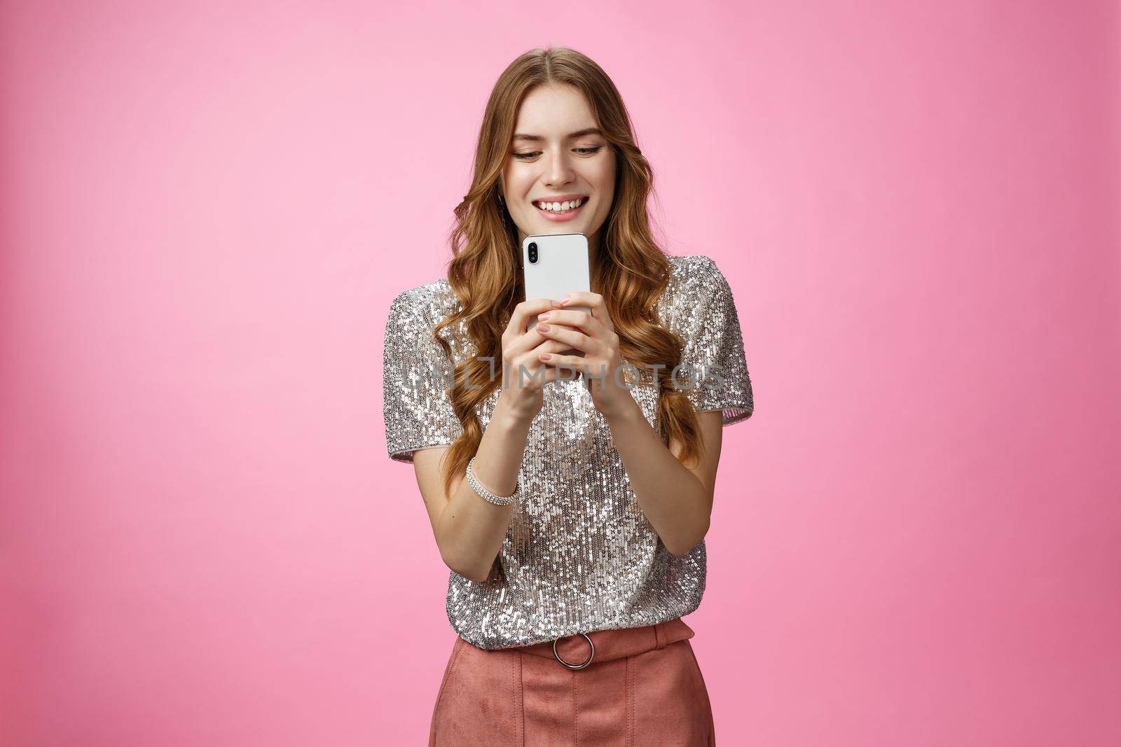 Charming glamour sociable stylish caucasian female using smartphone looking mobile phone screen look gadget display amused writing post upload photo social web page texting, pink background.