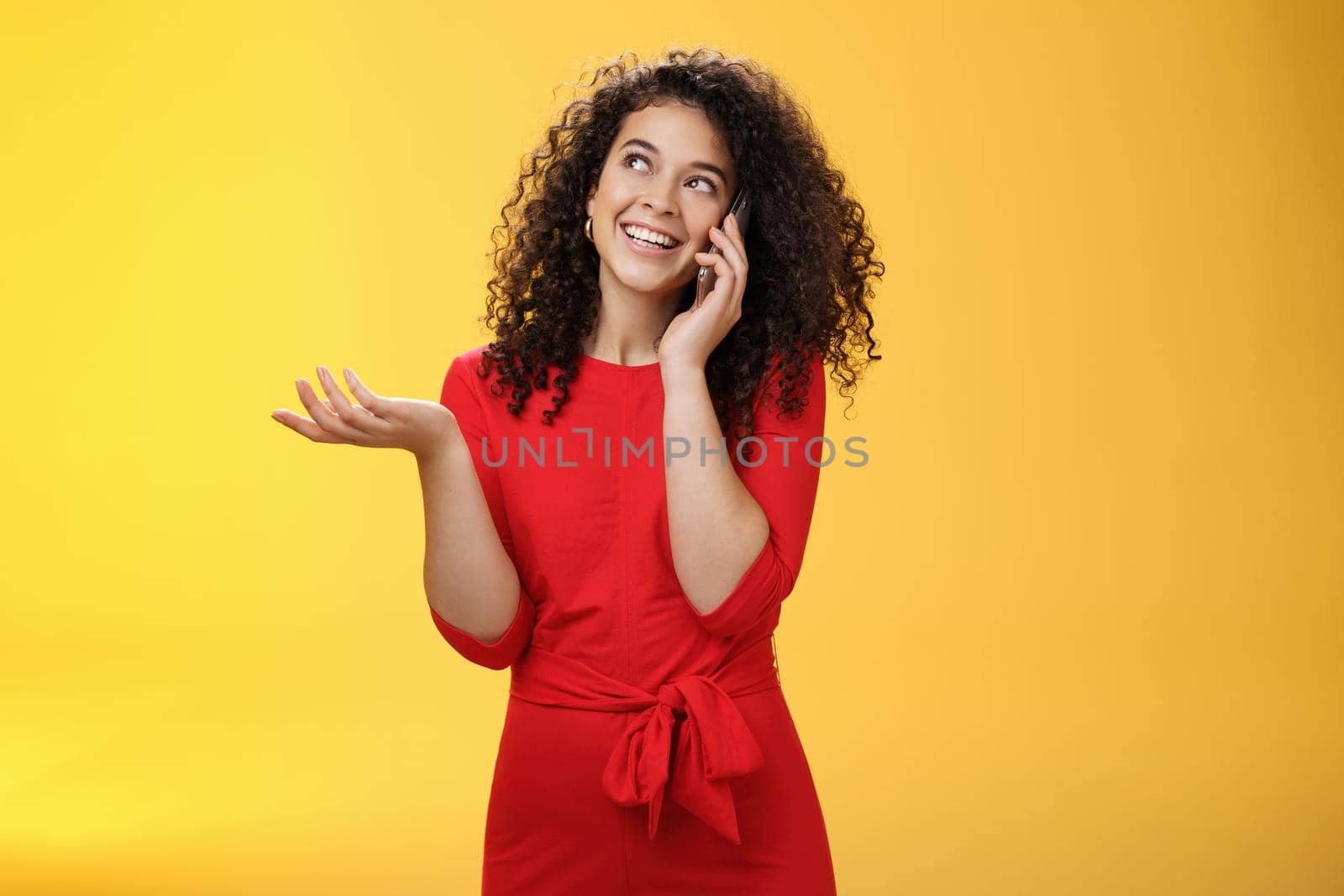 Carefree talkative cute woman with curly hair in red dress having char with friend on mobile phone holding smartphone near ear, gesturing as discussing exciting news, looking at upper left corner by Benzoix