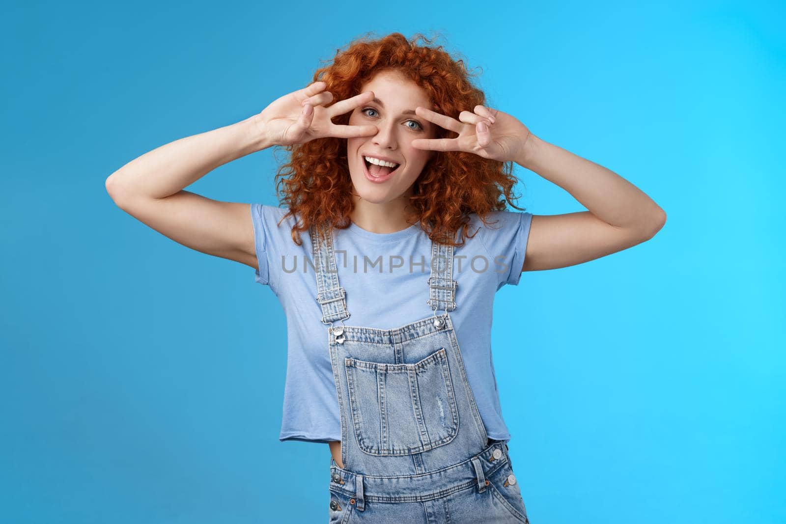 Cheerful cute redhead ginger girl curly haircut show positivity peace victory signs eyes smiling broadly have fun playful silly childish mood wear denim summer overalls blue background.